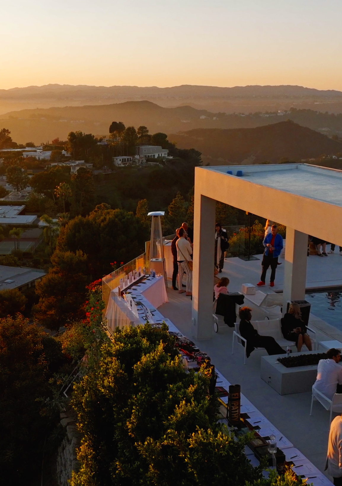 People gather on a rooftop terrace during sunset, with tables, chairs, and a pool, overlooking a hilly landscape with trees and residential buildings.