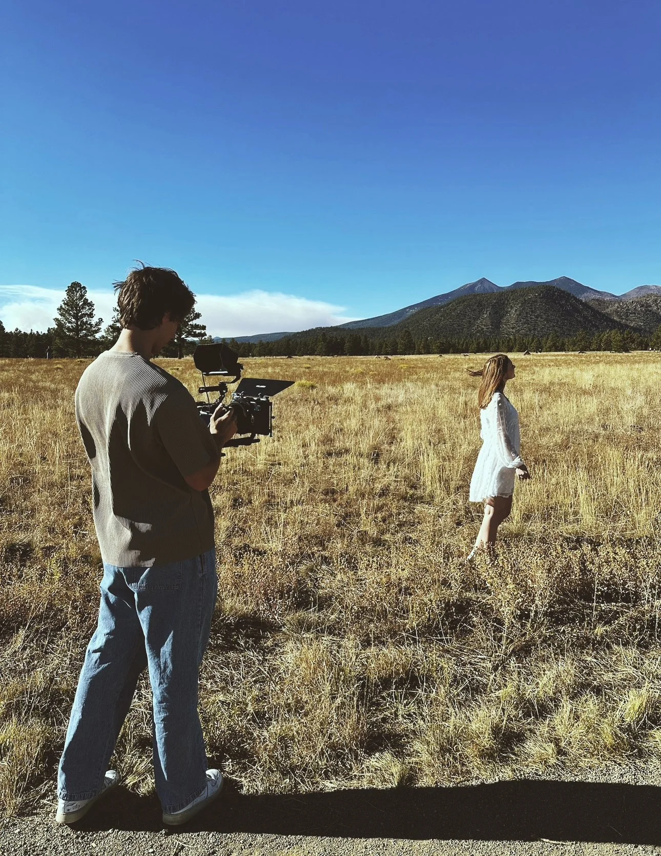 A person filming a woman standing in a field with mountains in the background on a bright, sunny day.