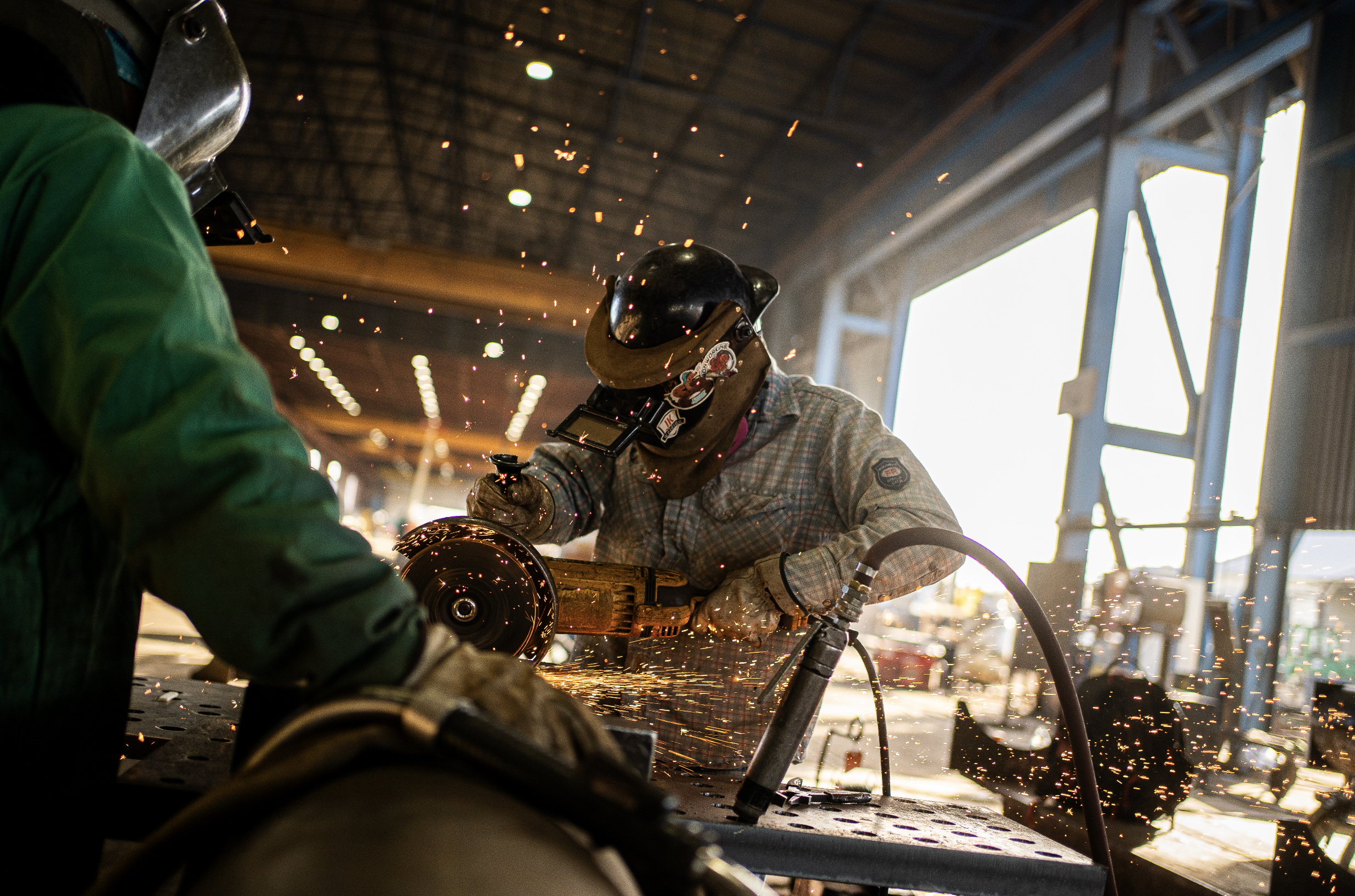 Welders in a large industrial workshop welding metal, with sparks flying during daylight.