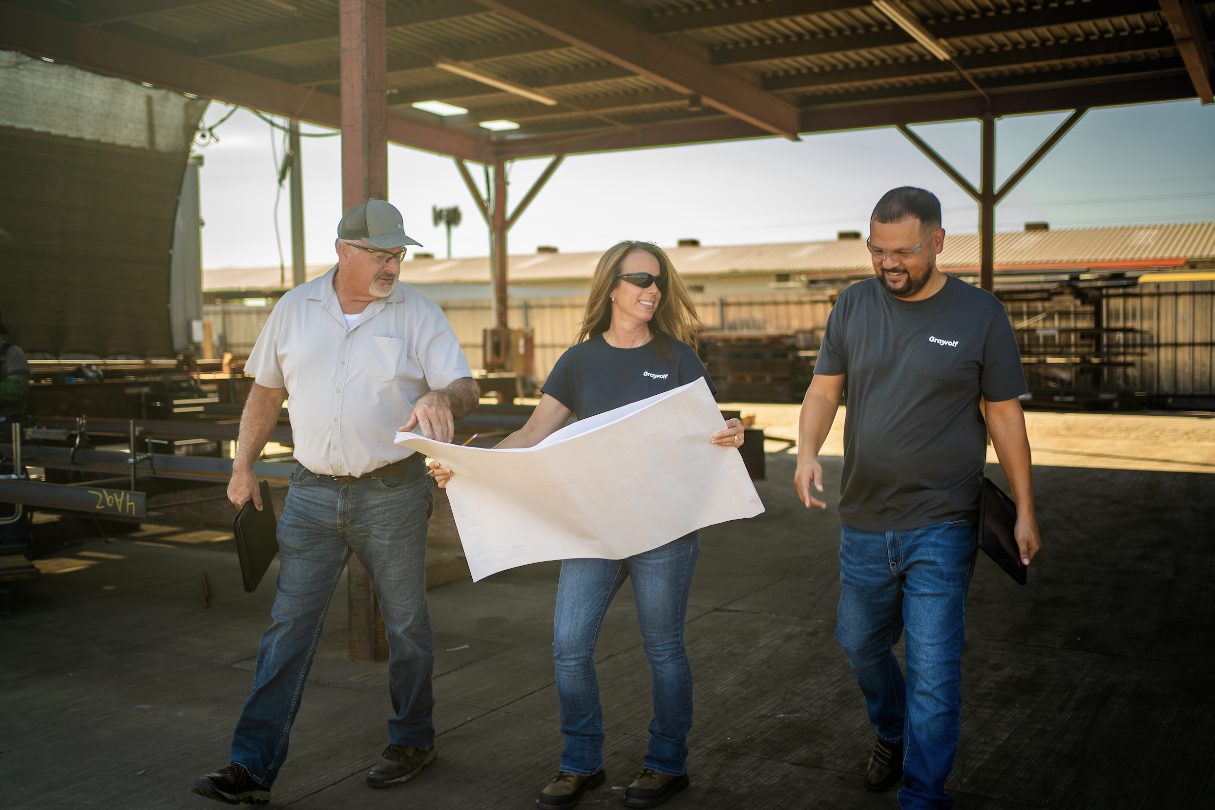 Three people walking and discussing blueprints in an outdoor covered area, with structures in the background.