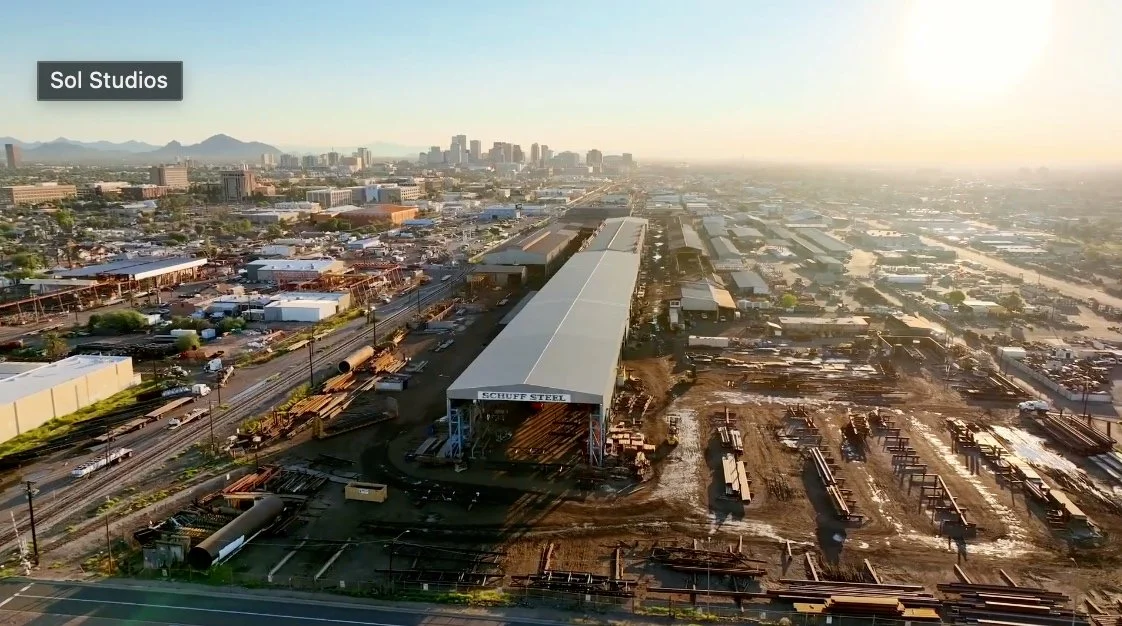 Aerial view of a steel manufacturing yard with a large building labeled 'Schuff Steel' and several steel beams and pipes stored on the ground, with a city skyline and mountains in the background during sunset.