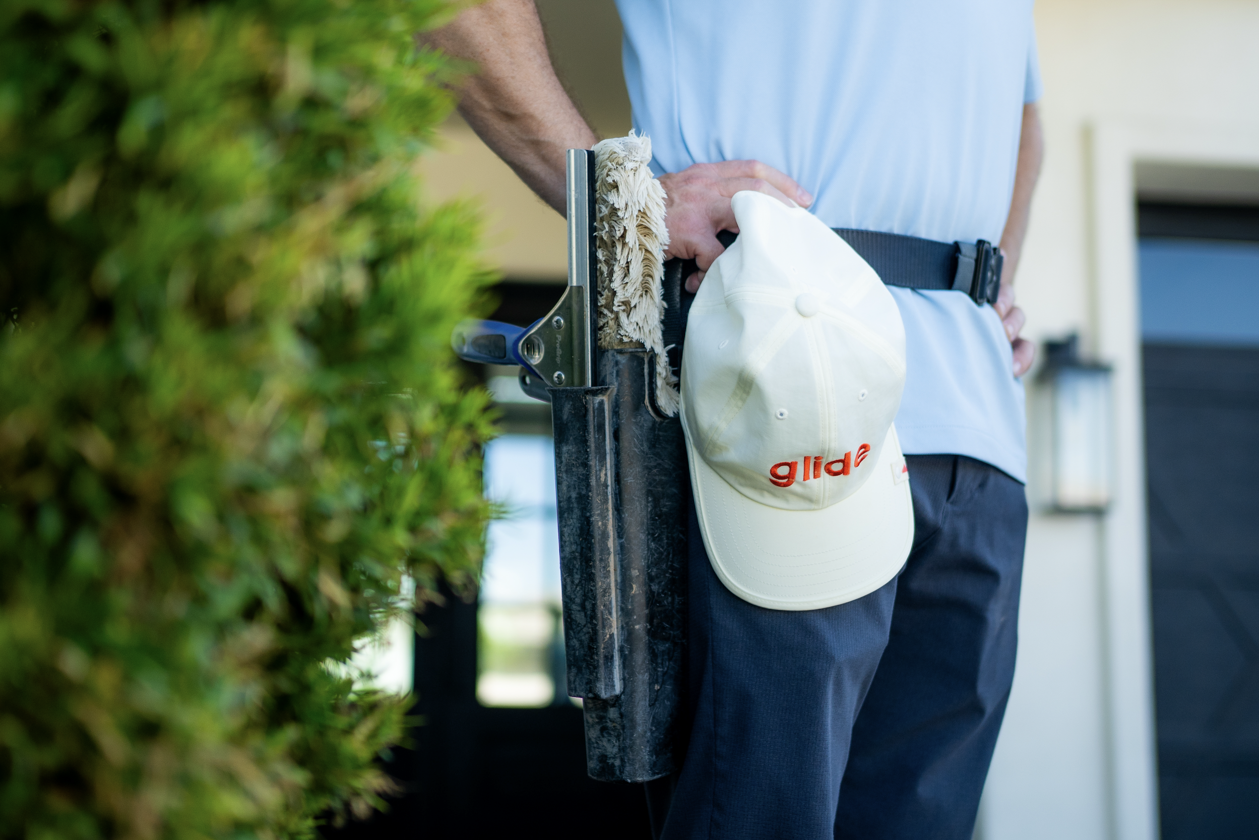 A person wearing a white glove holding a string trimmer with a cleaning cloth attached, a white cap hanging from the tool, and a light blue shirt, standing outside near green bushes.