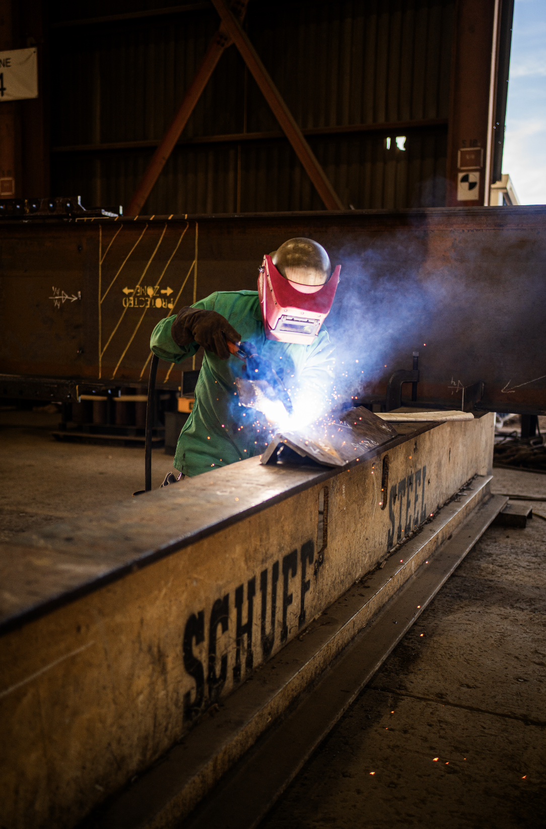 Welder wearing a red protective helmet and green uniform welding a metal beam inside a workshop.
