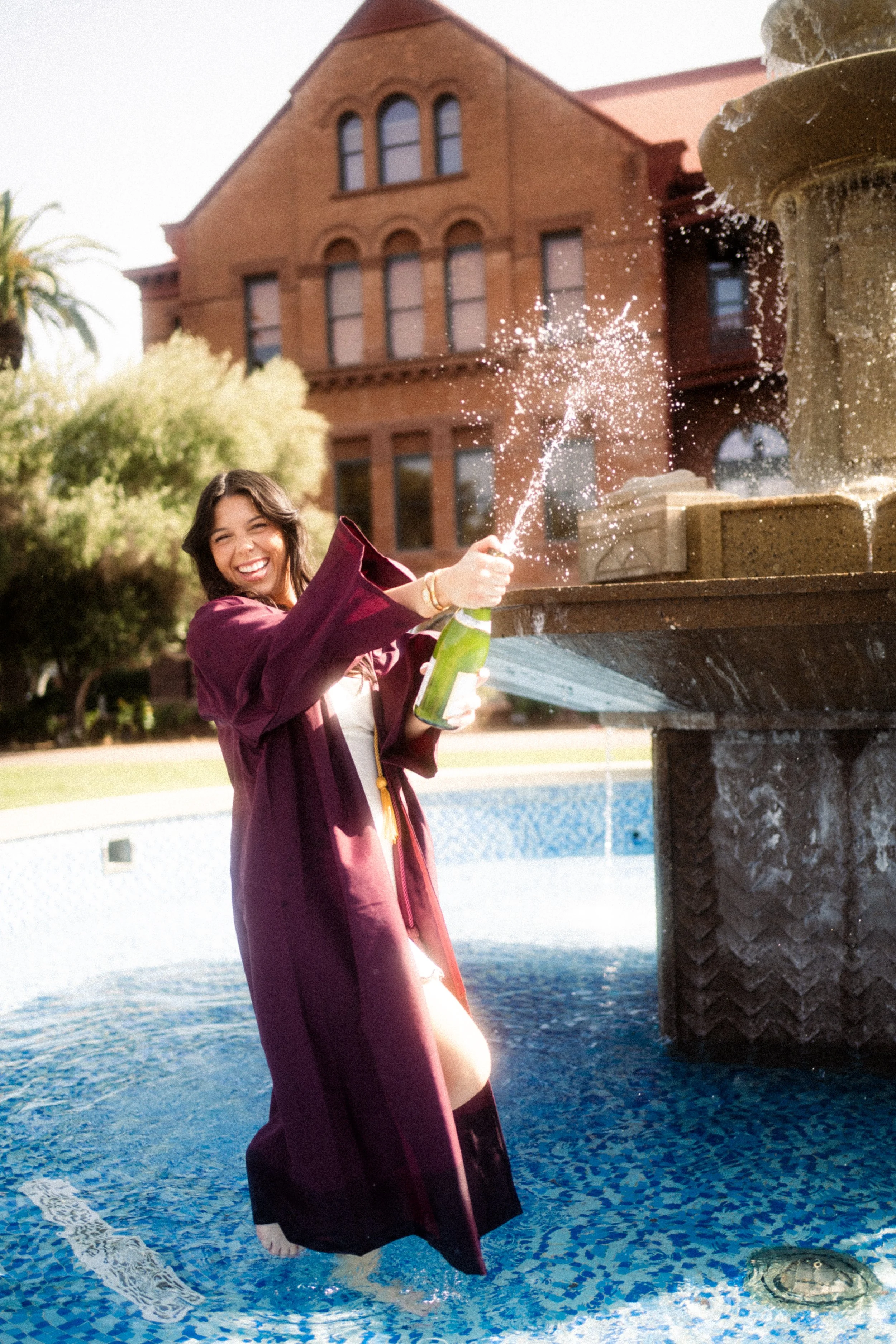 A young woman in a graduation gown celebrating with a bottle of champagne in front of a fountain.