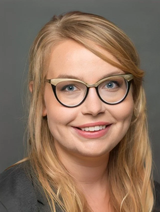 A woman with blonde hair and blue eyes wearing glasses and a dark top, smiling against a gray background.