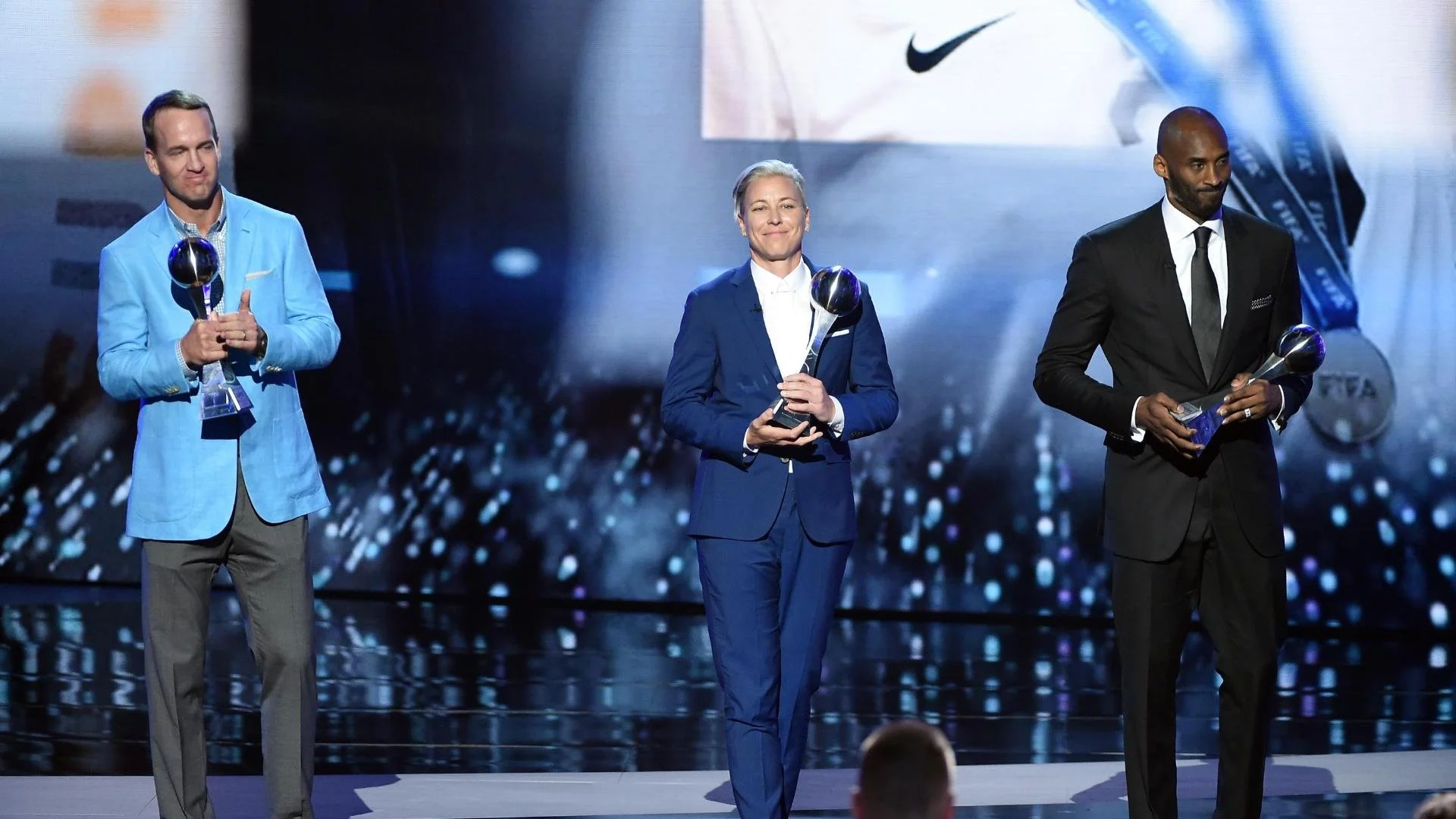 Three individuals on stage at an awards ceremony, holding trophies. The background is a large screen displaying images and logos.