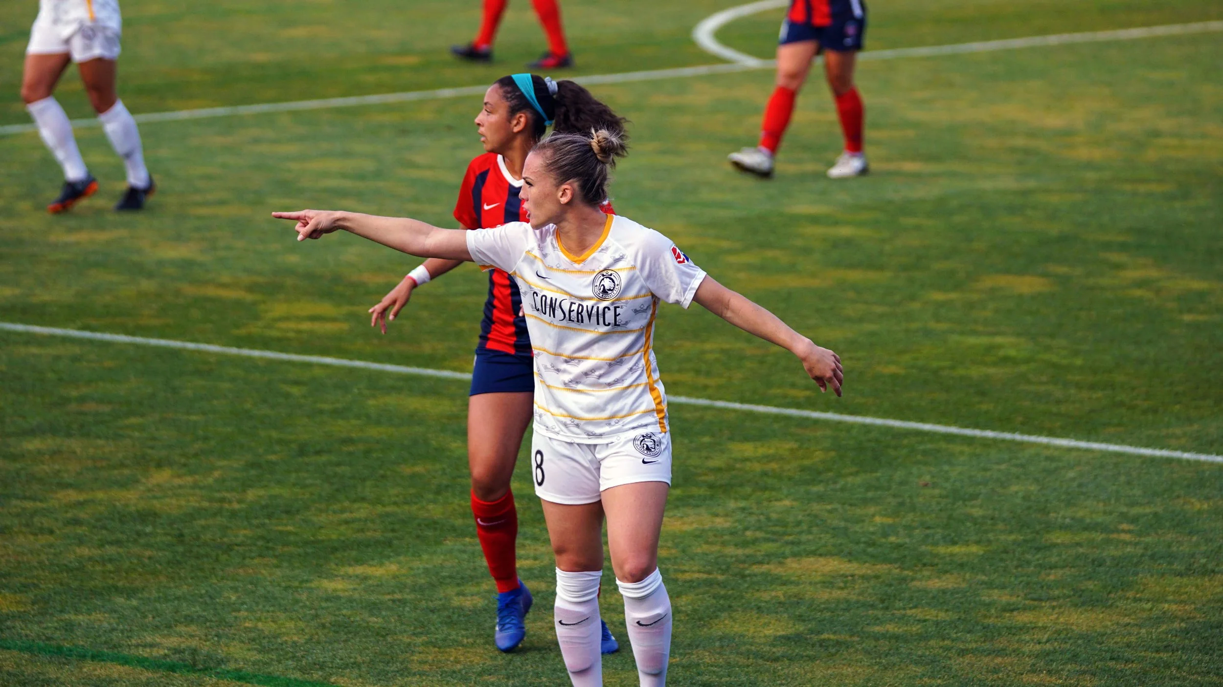 Soccer players on a field, one in a white jersey with gold accents pointing while another in a red and blue jersey stands nearby.