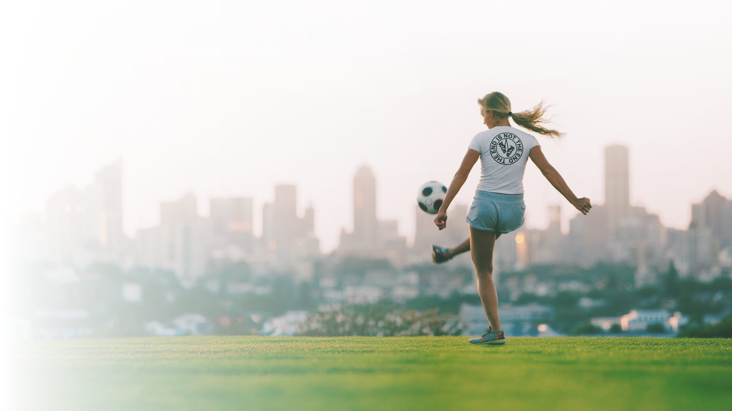 A young woman playing soccer on a grassy field with a city skyline in the background during sunset or sunrise.