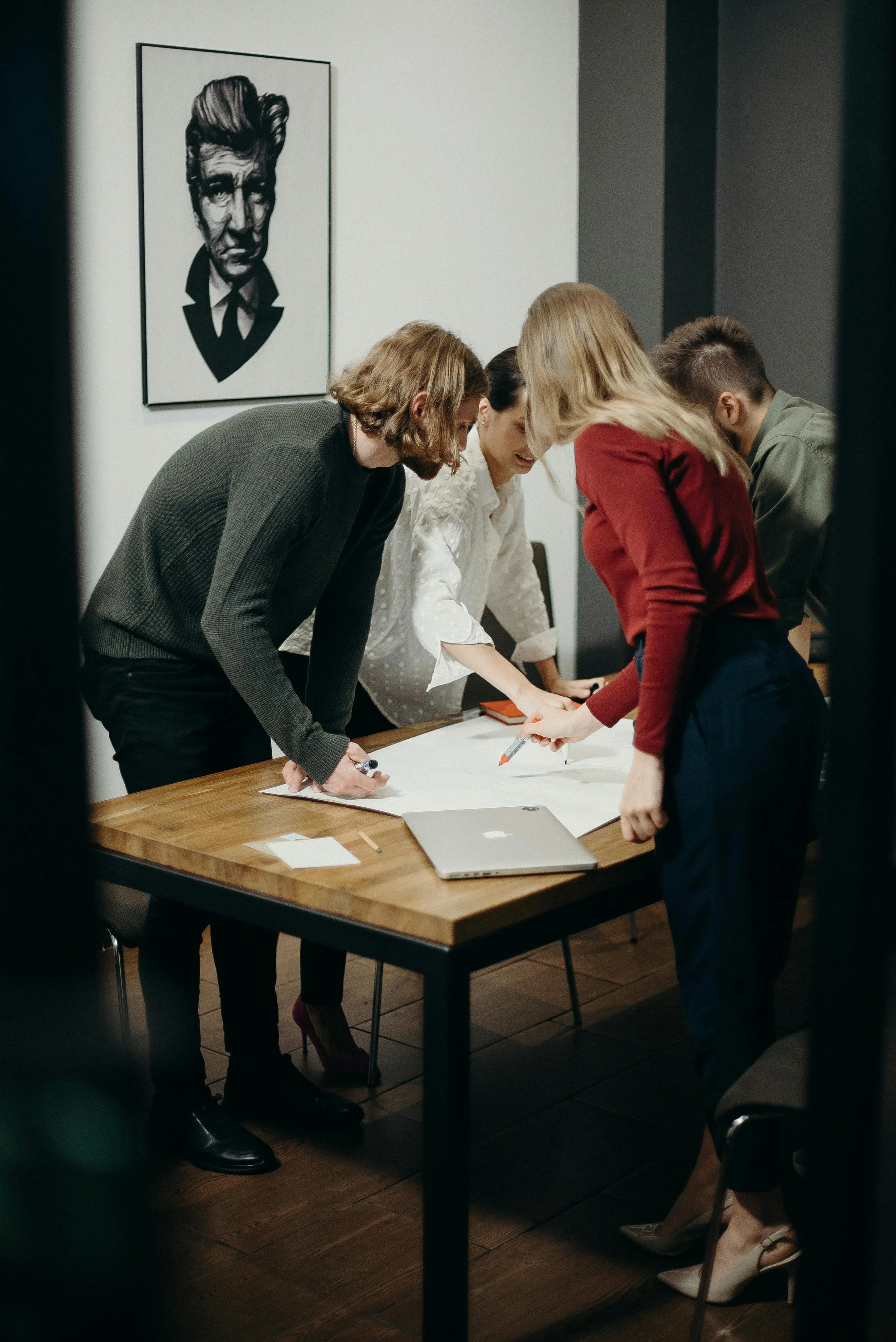 Four people gathered around a wooden table, reviewing a large sheet of paper with documents and a laptop on it, in a room with framed artwork on the wall.