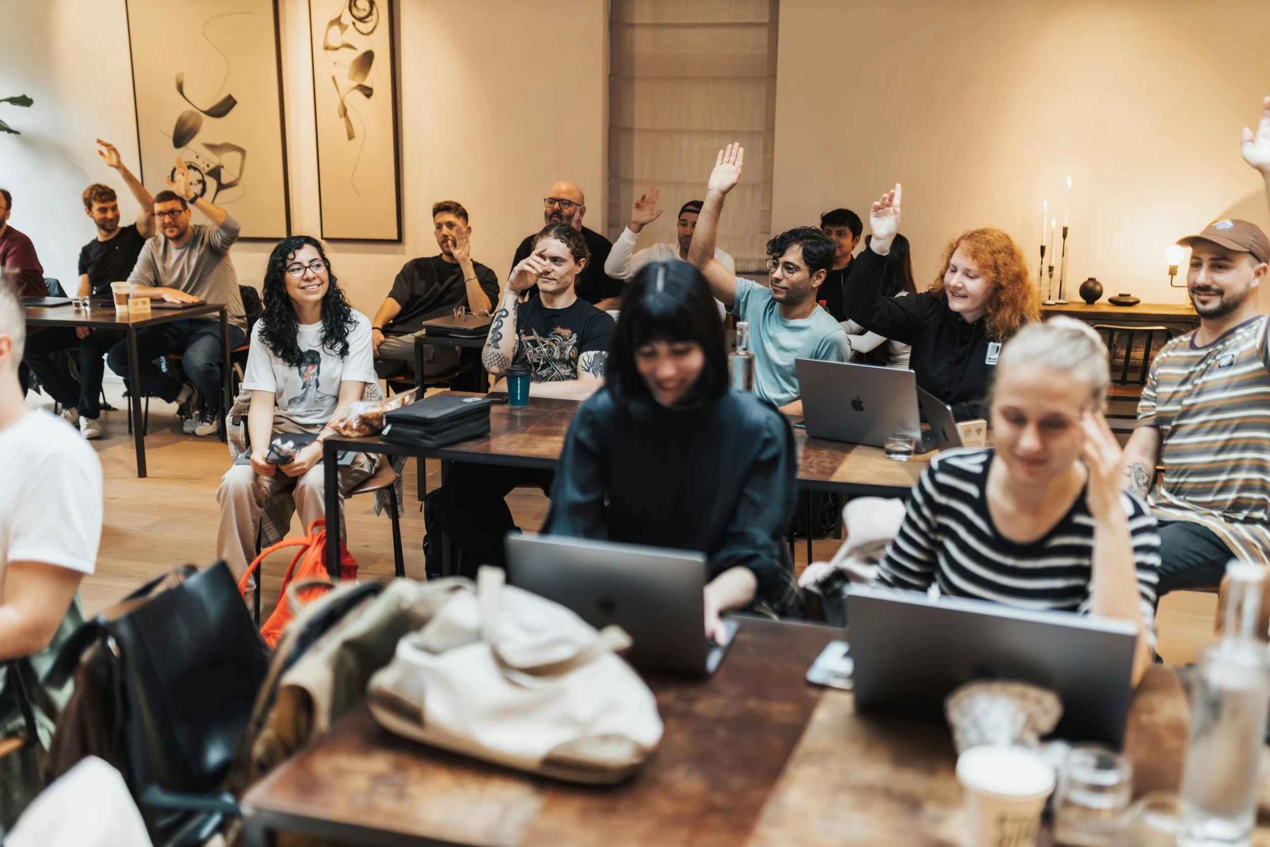 A group of diverse people in a meeting or workshop, some raising their hands, sitting at tables with laptops, notebooks, and drinks in a well-lit room with abstract wall art.