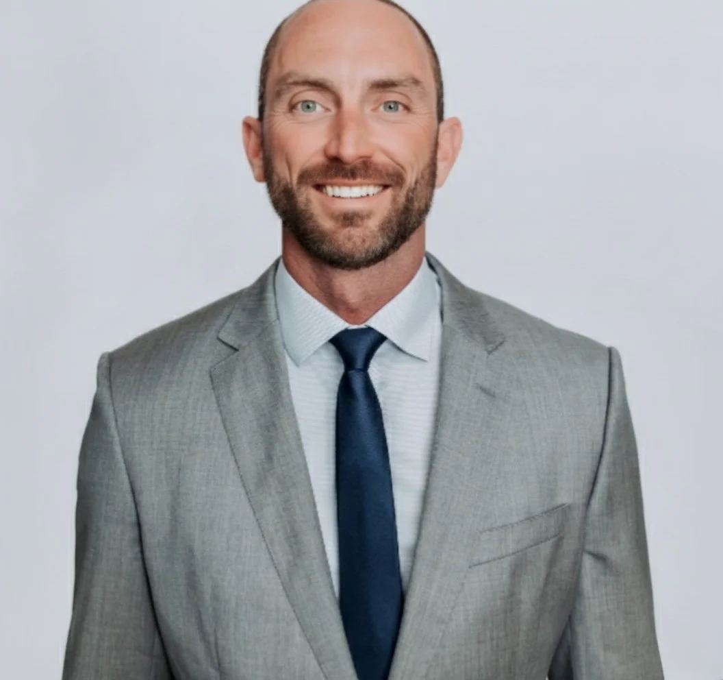 A smiling man in a light gray suit, white dress shirt, and navy tie, against a plain light background.