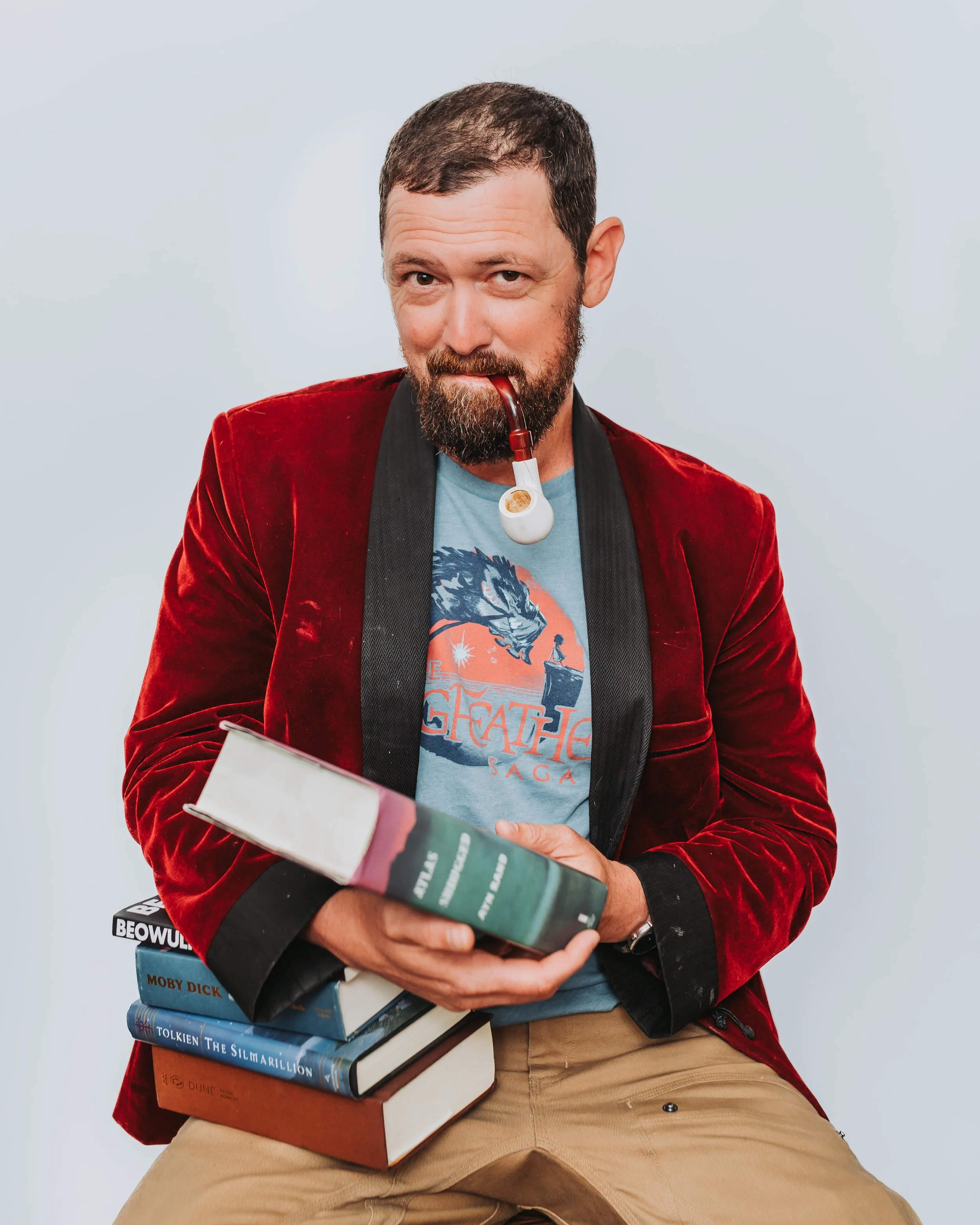 A man with a beard and short hair, wearing a red velvet blazer, a graphic t-shirt, and khaki pants, is sitting and holding a stack of books. He has a pipe in his mouth and is looking at the camera with a playful expression.