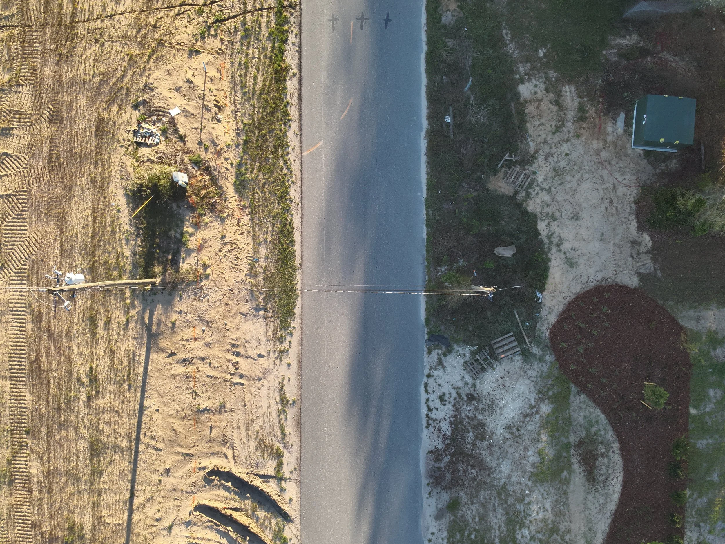 An aerial view of a roadside area with utility poles, some equipment, a patch of dark mulch, dirt, and a small green container, with sparse vegetation and tire tracks in the dirt.