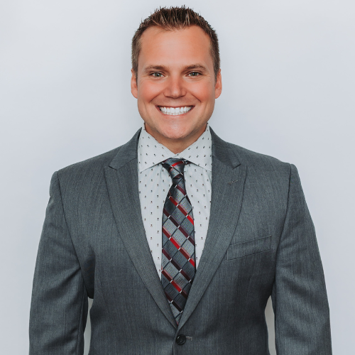 Smartly dressed man in gray suit with patterned tie smiling against a plain background.