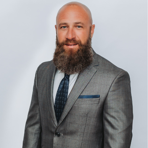 A man with a beard wearing a gray suit, white shirt, and dark tie, standing against a plain light background.