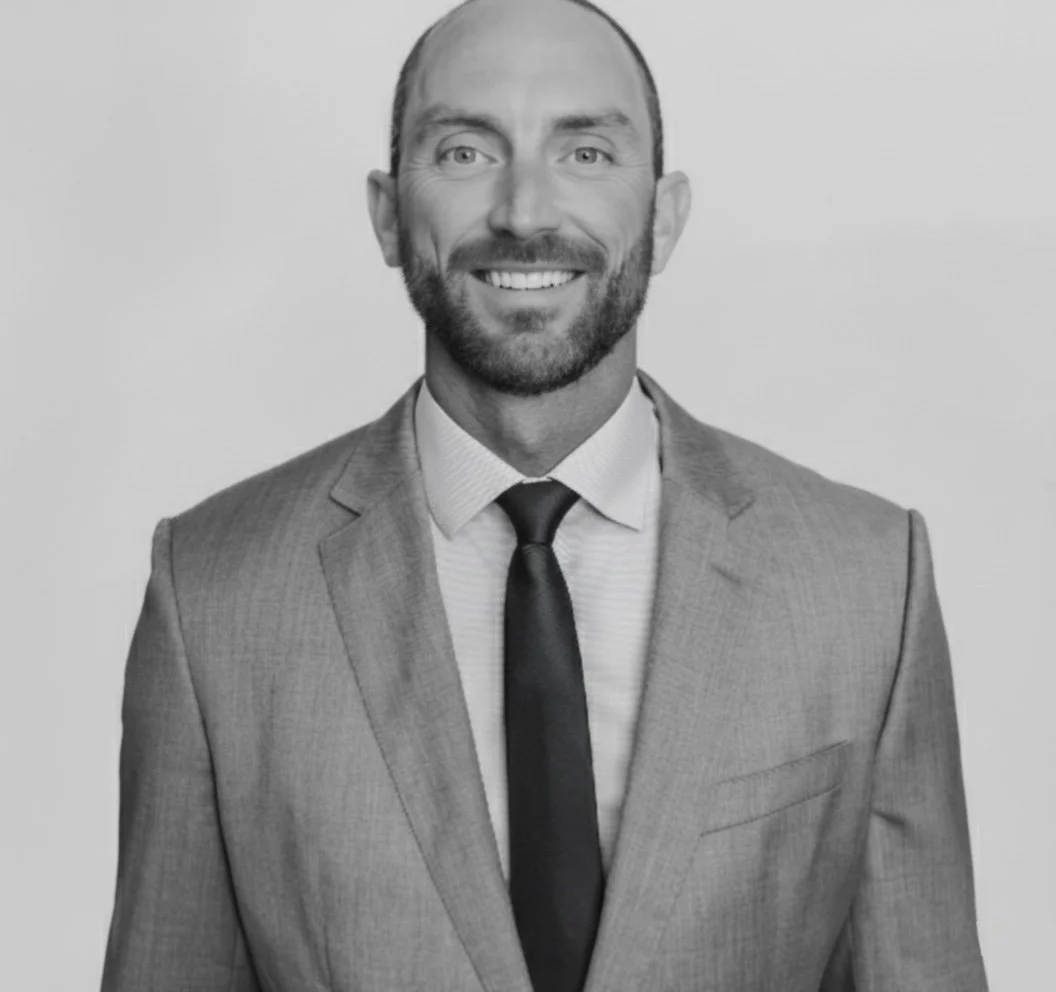 A smiling man in a light-colored suit with a tie, standing against a plain background.