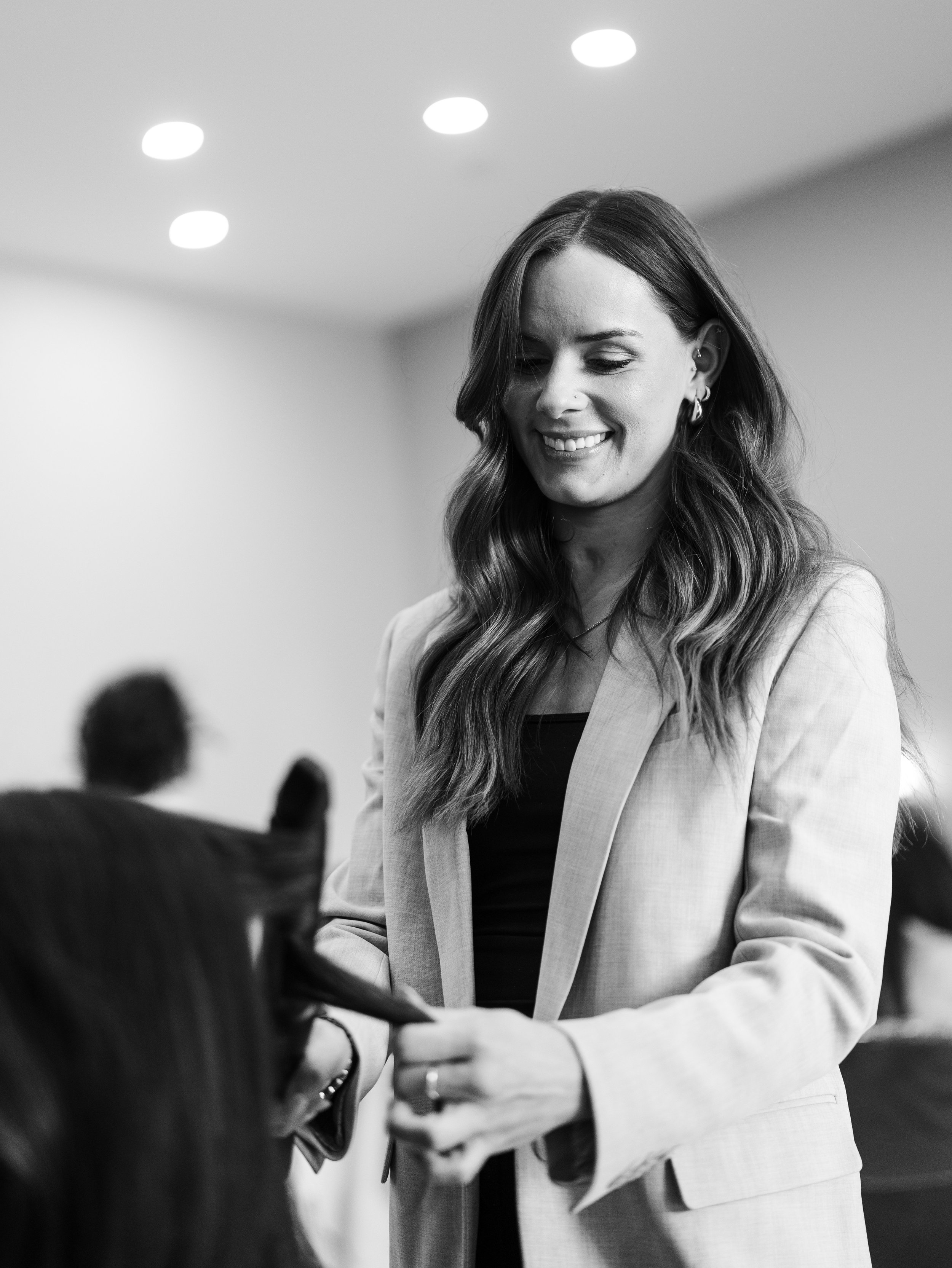 A smiling woman with long wavy hair and earrings in a light-colored blazer interacting with someone out of frame.