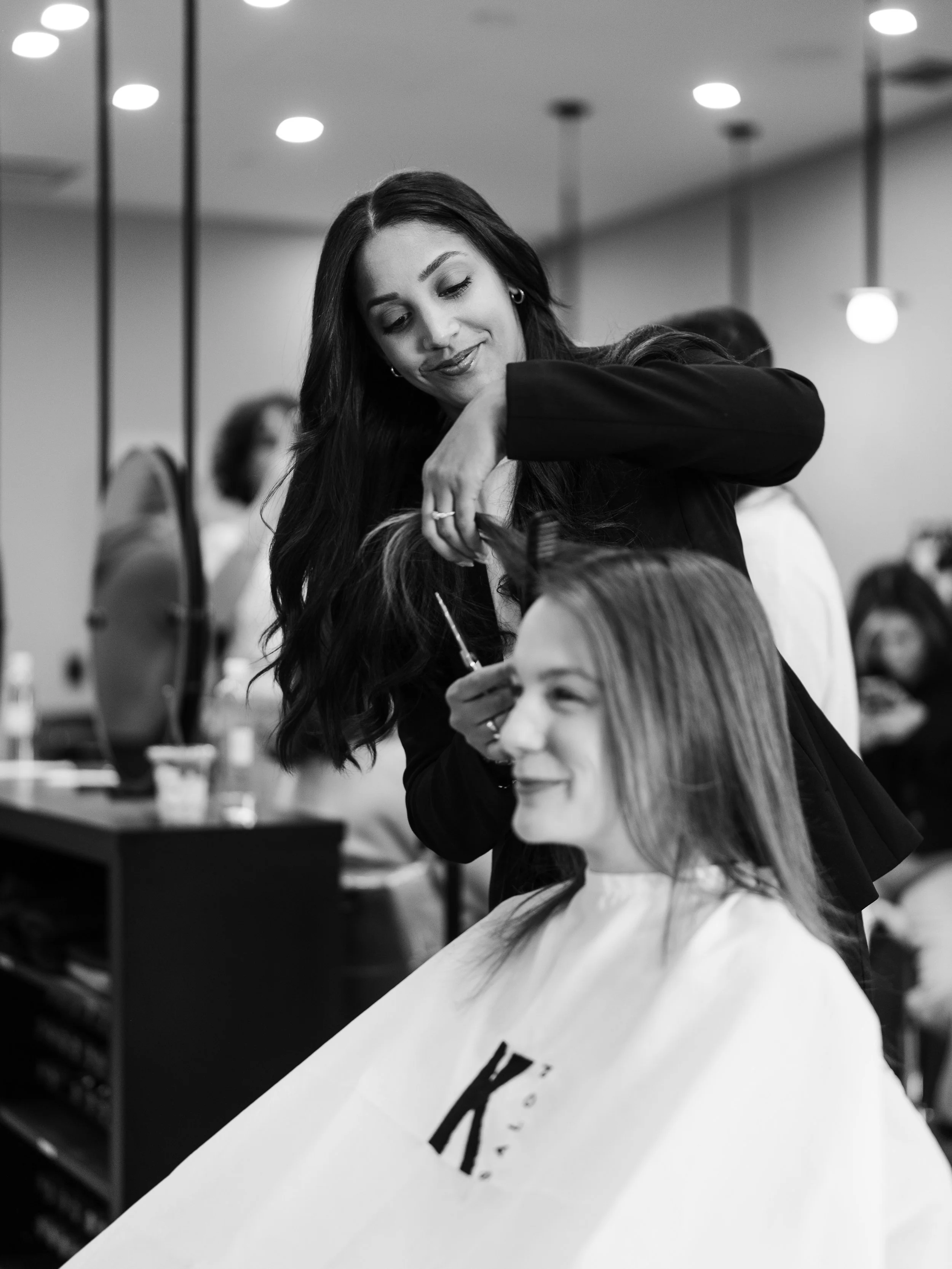 A woman with long dark hair is styling another woman's hair at a salon or beauty parlor, with salon equipment and other clients in the background.