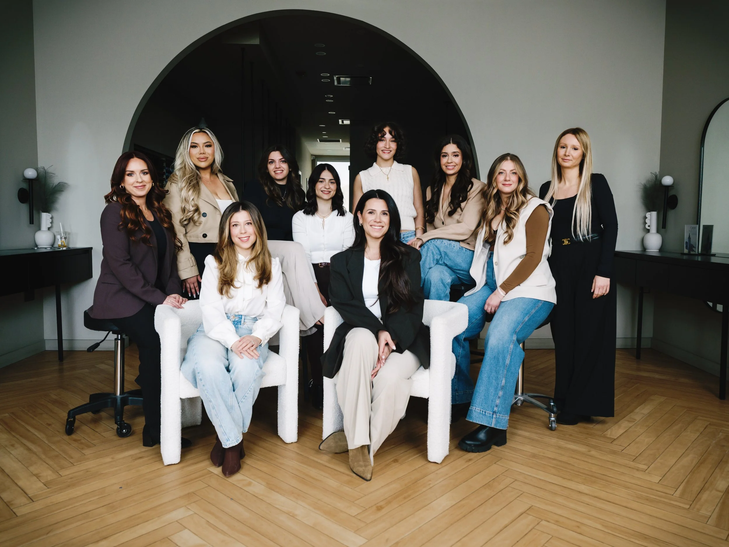 A group of twelve diverse women in casual and professional attire posing together in a modern interior space with hardwood floors, black and white furniture, and decorative lighting.