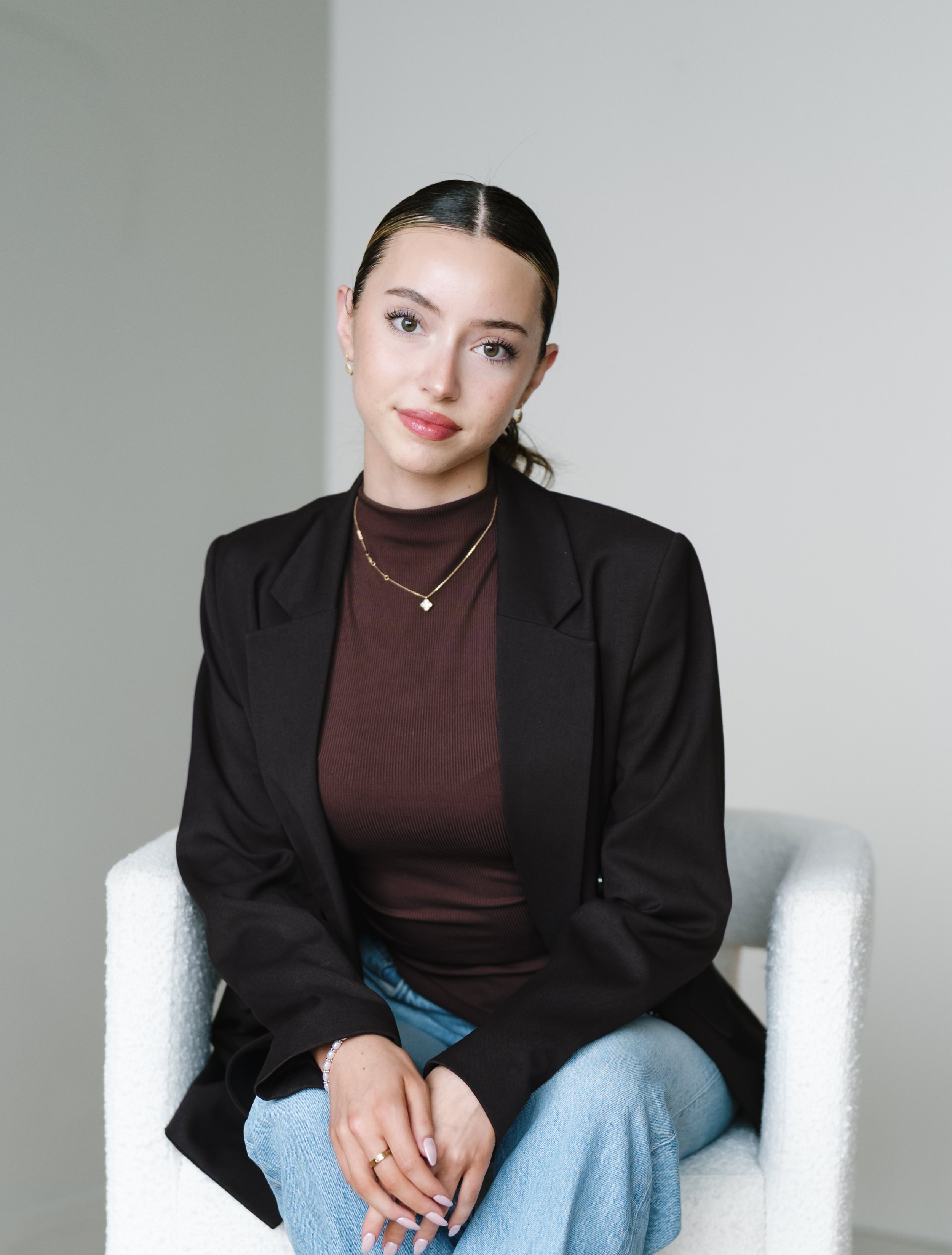 A young woman with dark hair, light skin, and green eyes sitting on a white armchair against a plain gray and white wall. She is wearing a black blazer, dark red top, layered necklaces, earrings, and rings, with her hands resting on her lap.