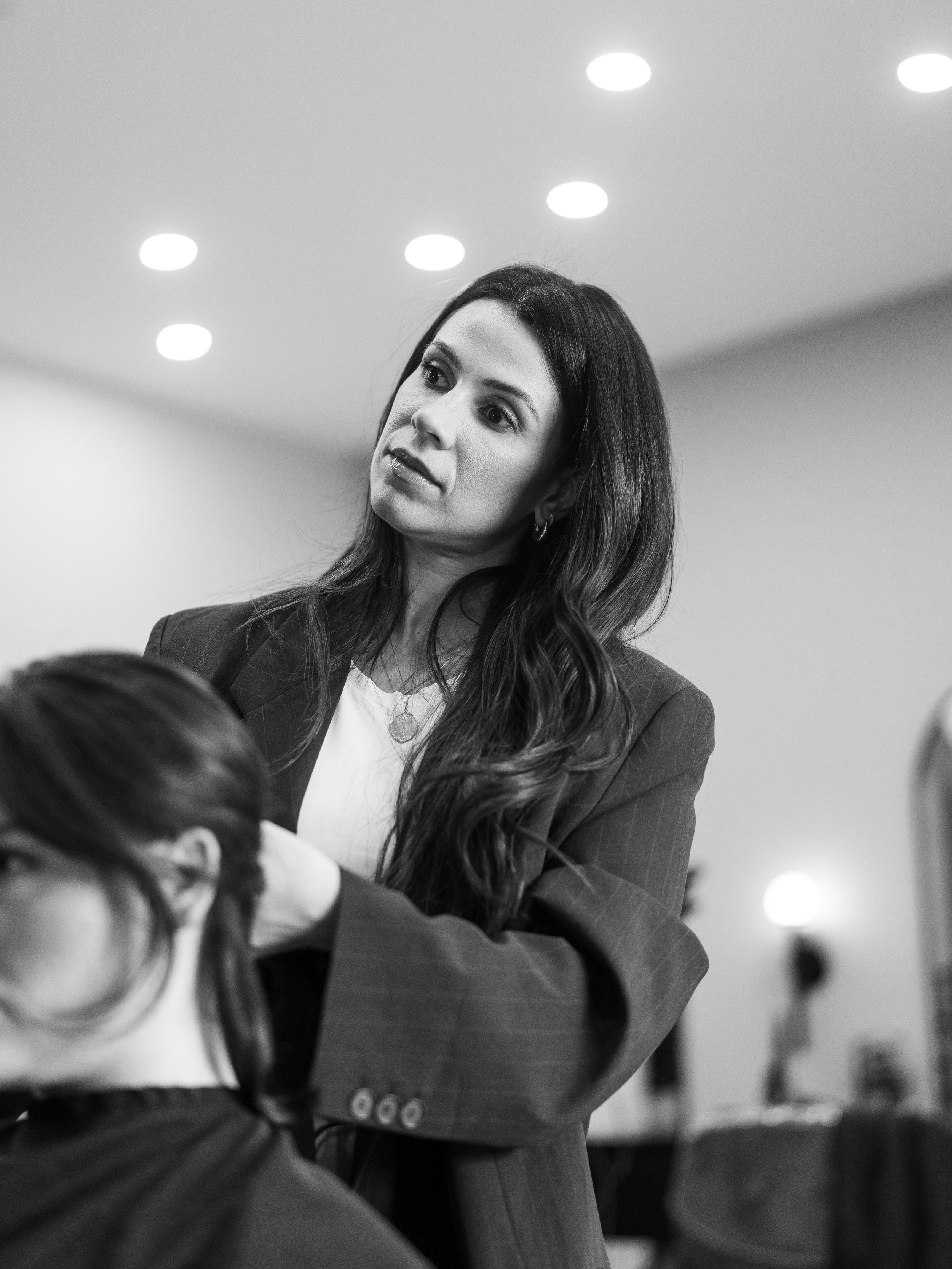 Black and white photo of a woman with long dark hair, wearing a striped suit, sitting at a table, looking slightly to her right. The ceiling with round lights is visible in the background.