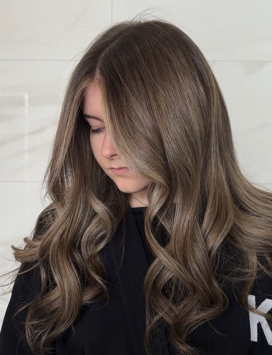 A woman with long, wavy, brunette hair looking downward, wearing a black top, against a white tiled background.