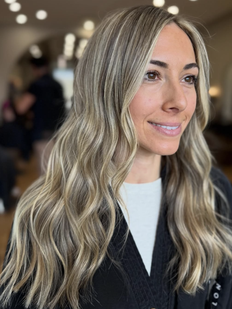 Close-up of a smiling woman with long, wavy blonde hair in a salon or similar setting.