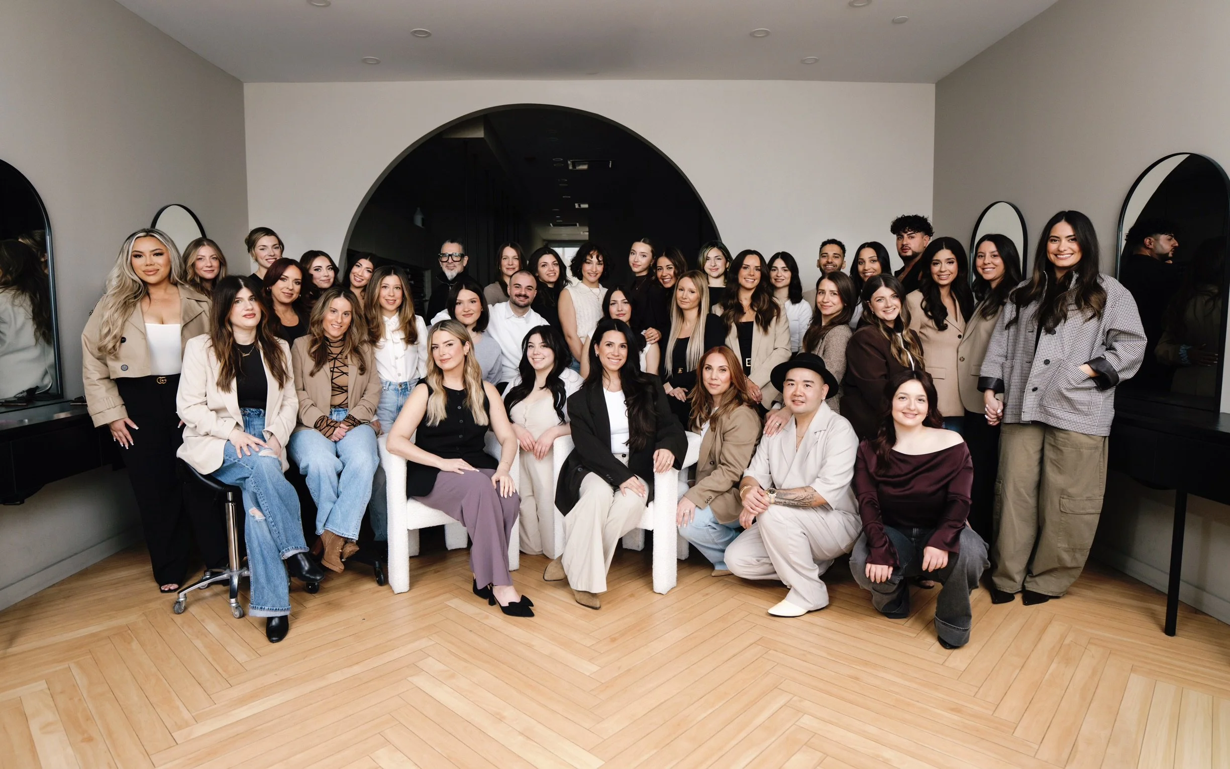 Large group of diverse women and a few men in semi-formal attire posing together in a room with wooden flooring and white walls, some smiling at the camera.