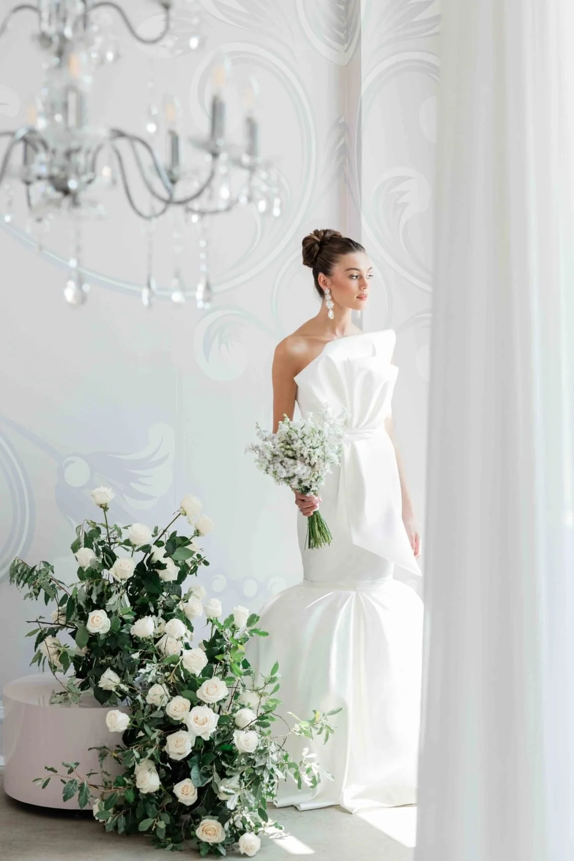 A bride in a white wedding dress holding a bouquet of white flowers during a wedding celebration indoors near a window, with a floral arrangement of white roses and greenery on a table, and a chandelier hanging from the ceiling.