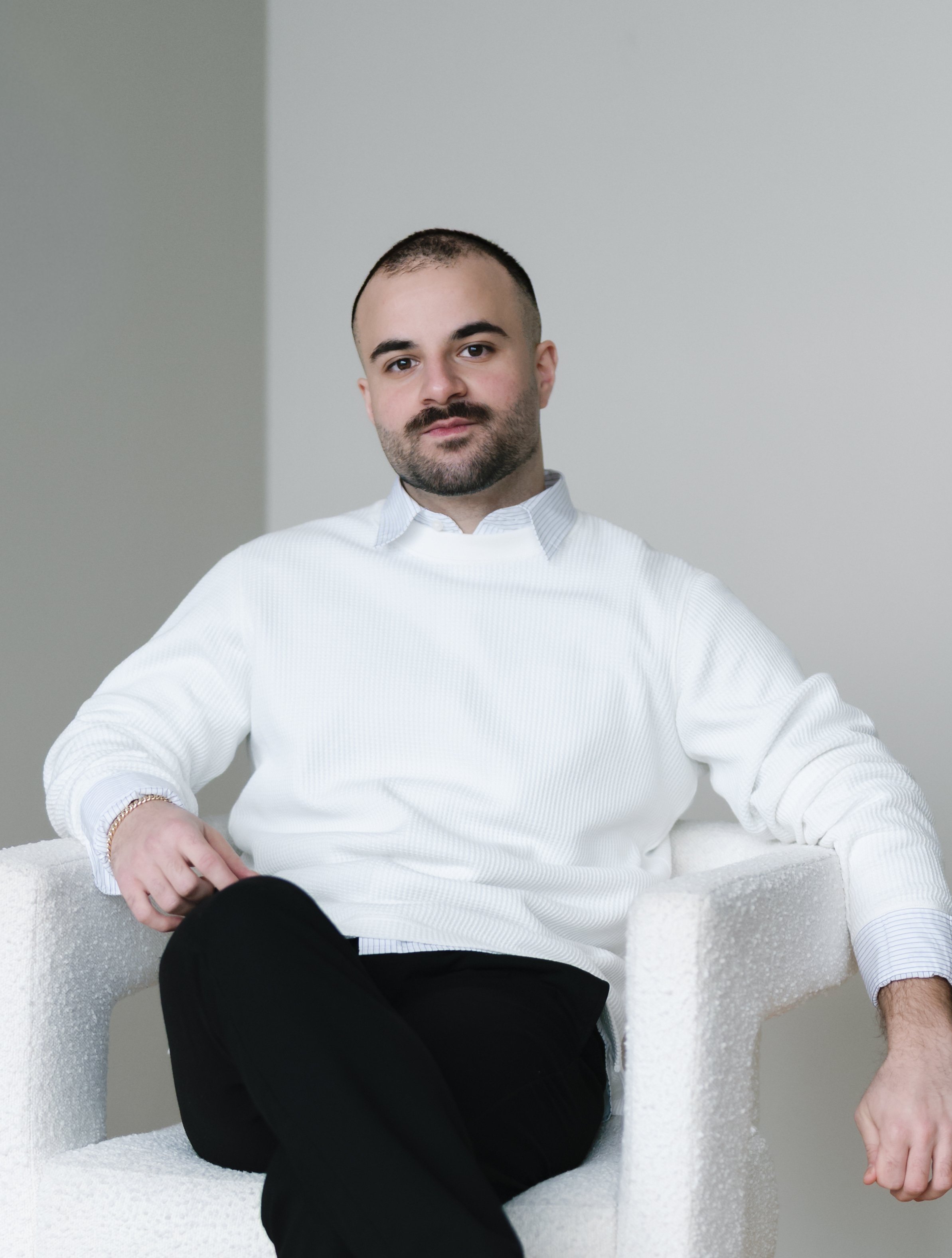 Black and white portrait of a young man with a beard, smiling, wearing a black button-up shirt, standing against a white background.