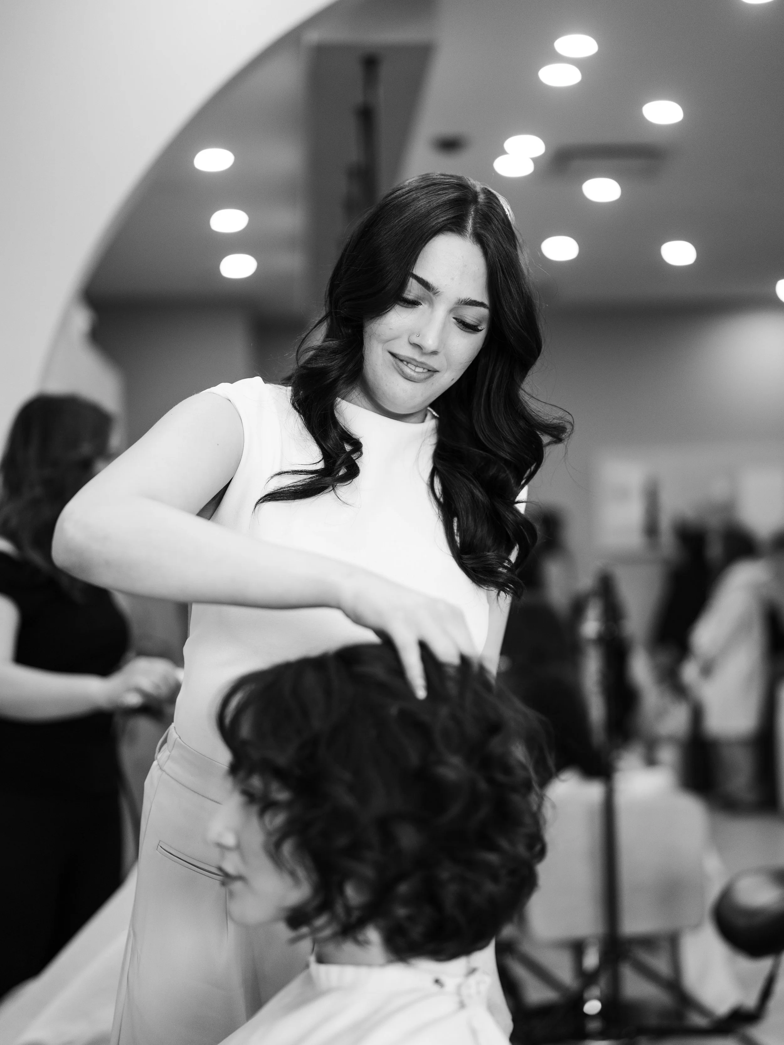 A woman with long dark hair styled in loose waves, wearing a white sleeveless top, is smiling and looking down while getting her hair done.