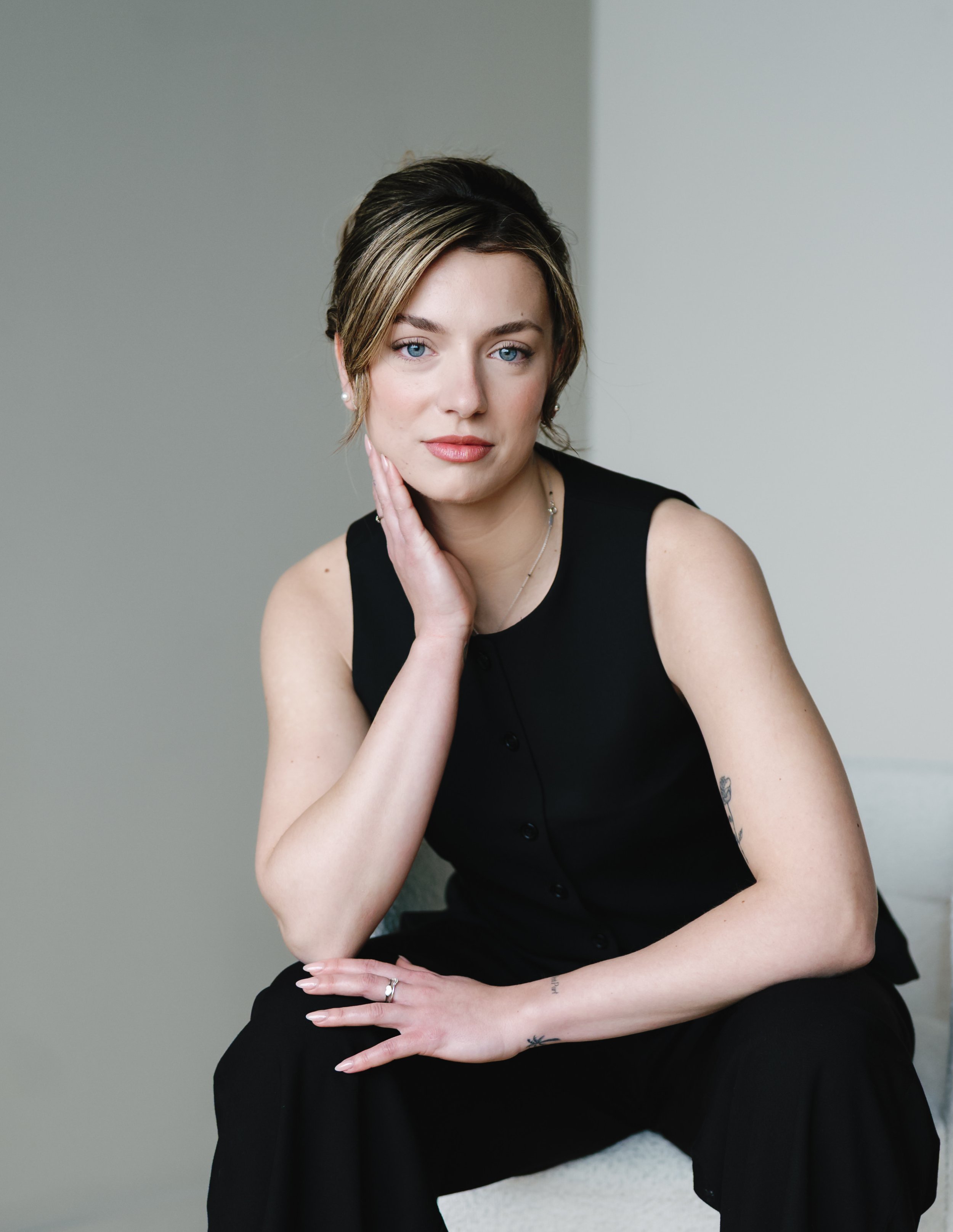 A woman with short, brown hair with blonde highlights, wearing a black sleeveless top, sitting against a plain wall, resting her right hand on her face and her left hand on her lap.