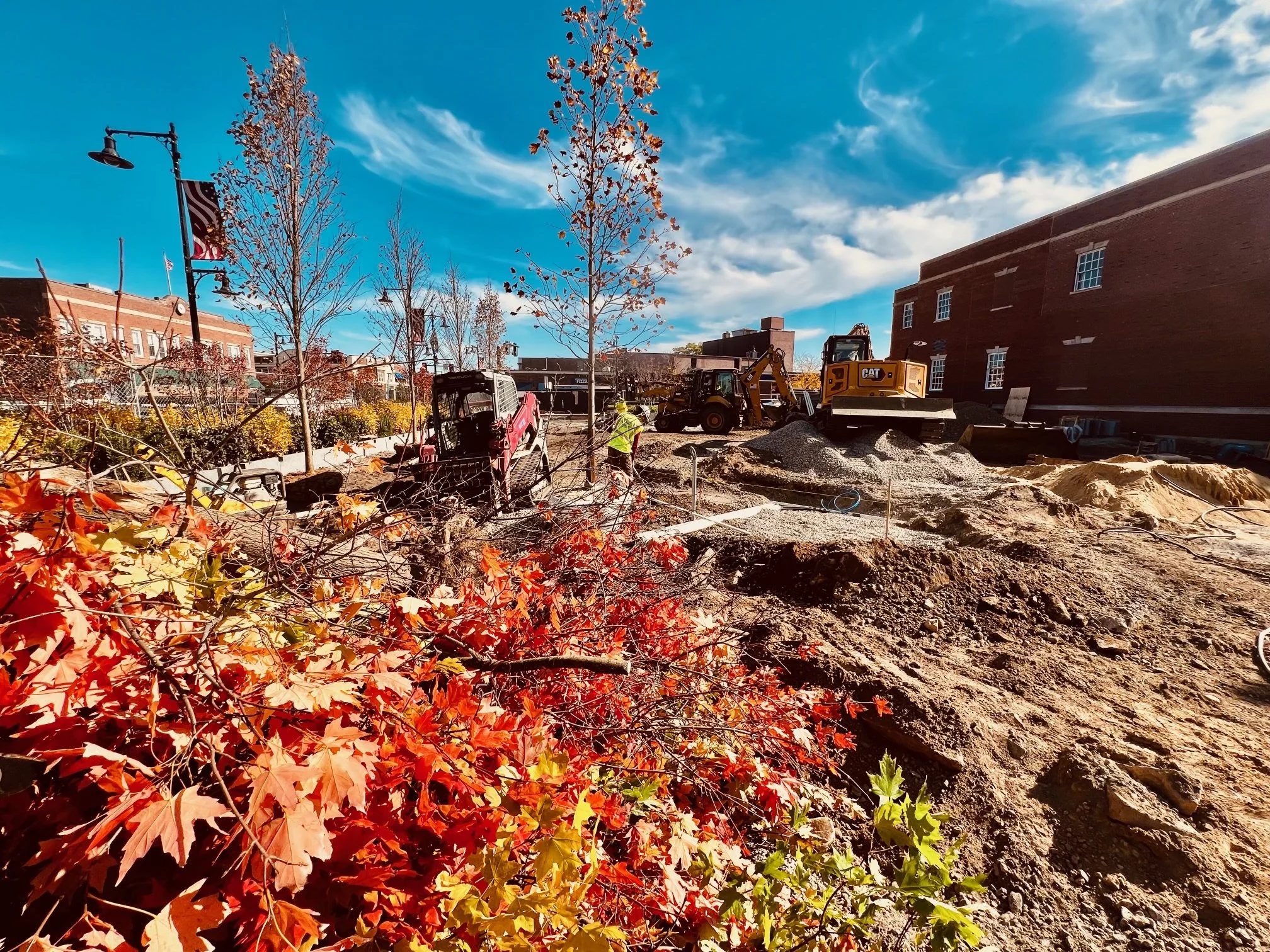 Construction site with heavy machinery and workers, autumn trees and a bright blue sky with clouds.