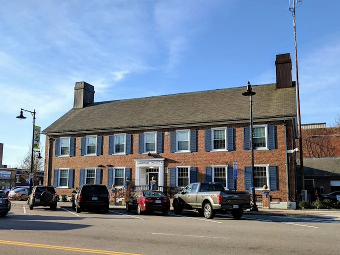 The former Dedham Police Station, a red brick building with blue shuttered windows and a white entrance, parked cars in front, clear blue sky overhead.