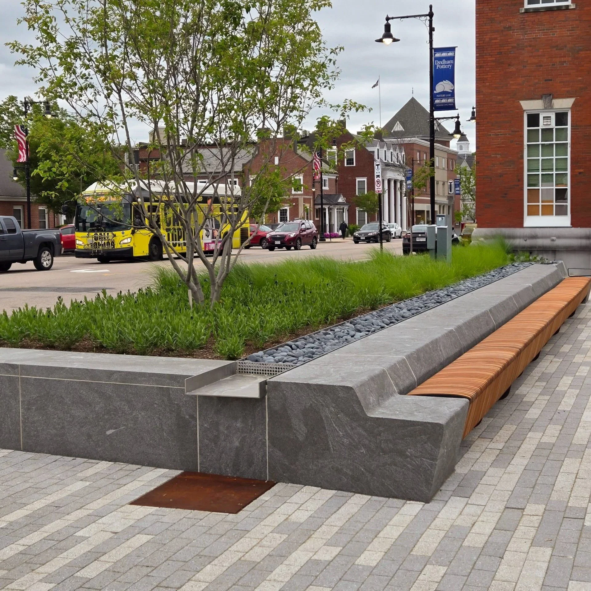 Granite benches next to a landscaped area with green grass, small shrubs, and a young tree. In the background, there's a street with parked cars, a yellow bus, and historic brick buildings, some with banners and American flags.