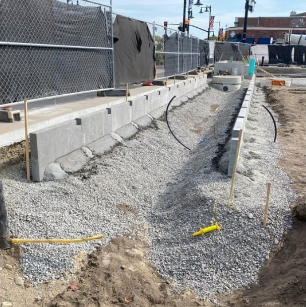 Construction site with a concrete curb, gravel, safety fencing, and construction tools along a city street.