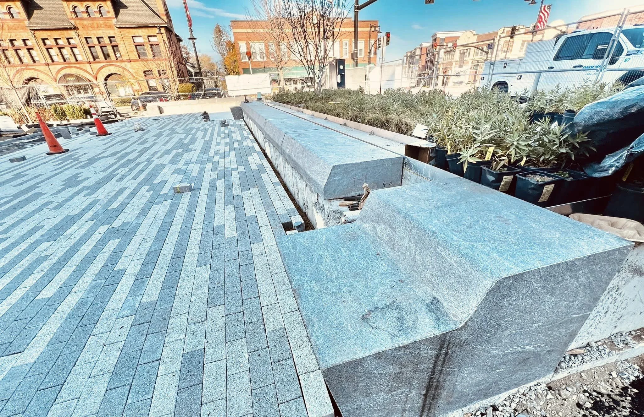 Close-up of a stone bench under construction with concrete blocks and pavers arranged for installation, with buildings, trees, and a street scene in the background.