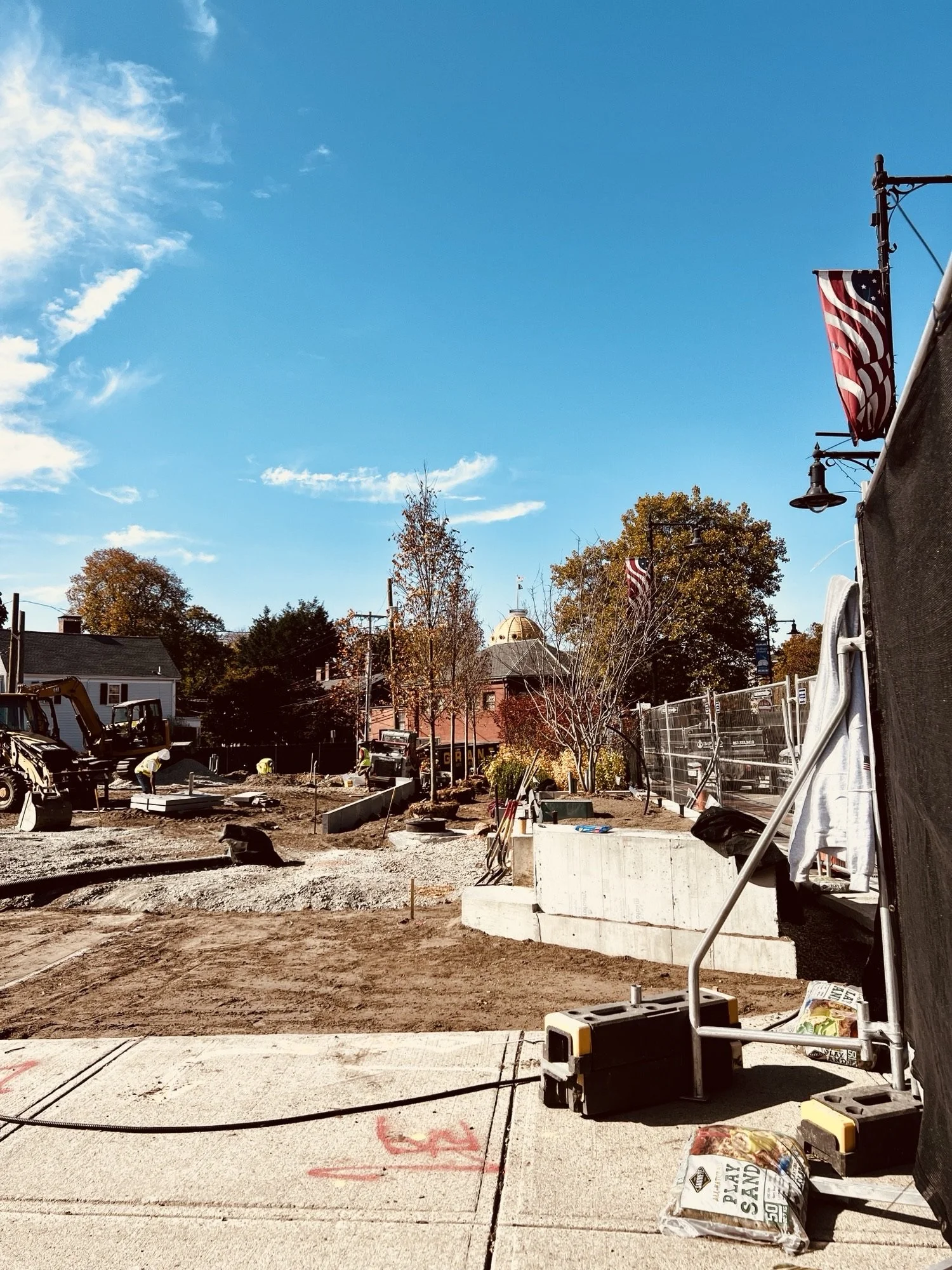 Town Green construction site with machinery, plants, and American flags on poles, under a clear blue sky with scattered clouds.