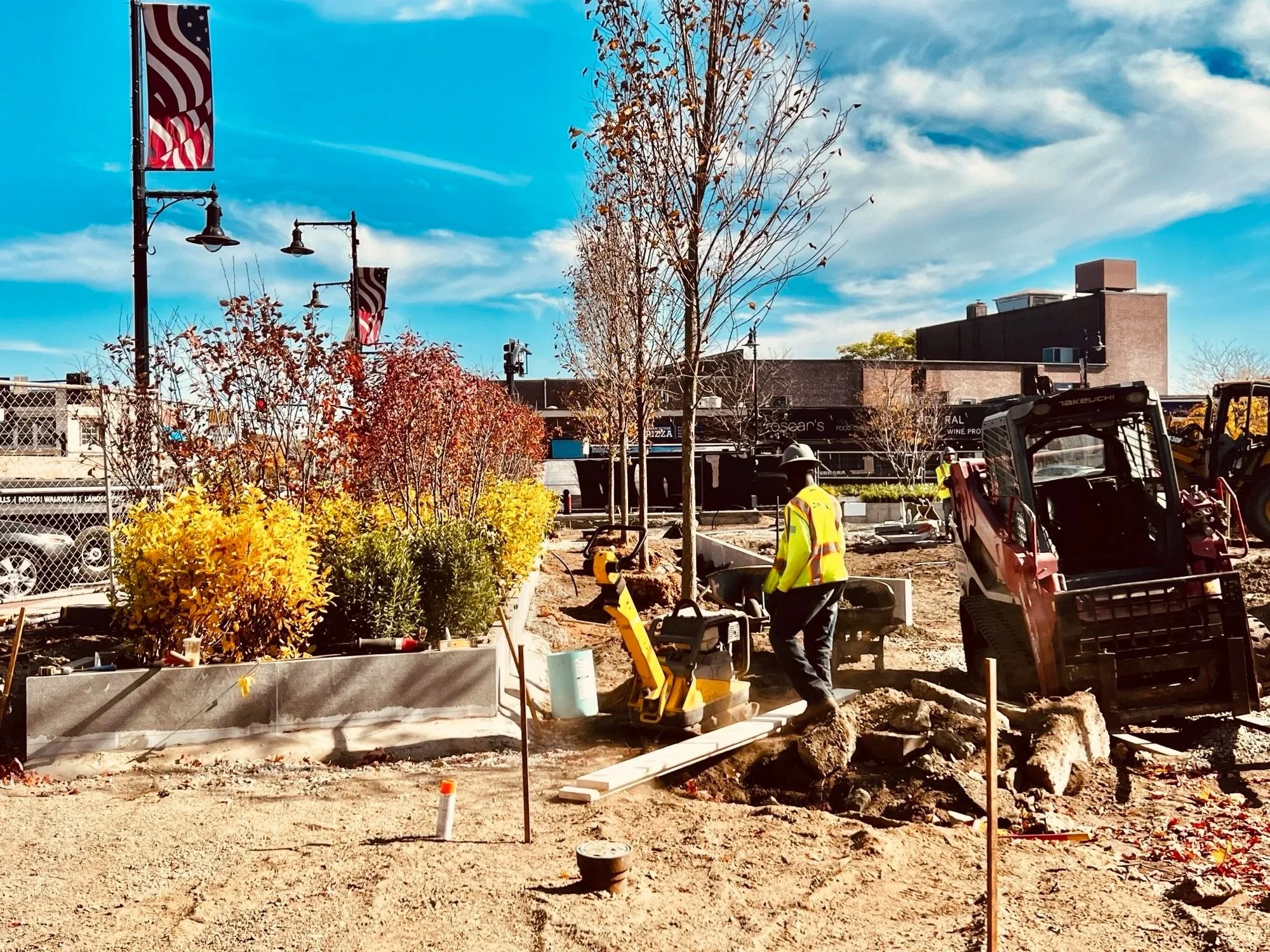 Construction worker operating a compactor machine at a landscaping site with decorative bushes, trees, and a cityscape background under a partly cloudy sky.