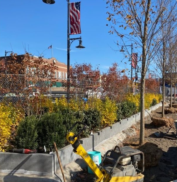 Town Green construction site with a garden with colorful bushes and small trees; American flags on flagpoles; construction tools and materials in the foreground; clear blue sky.