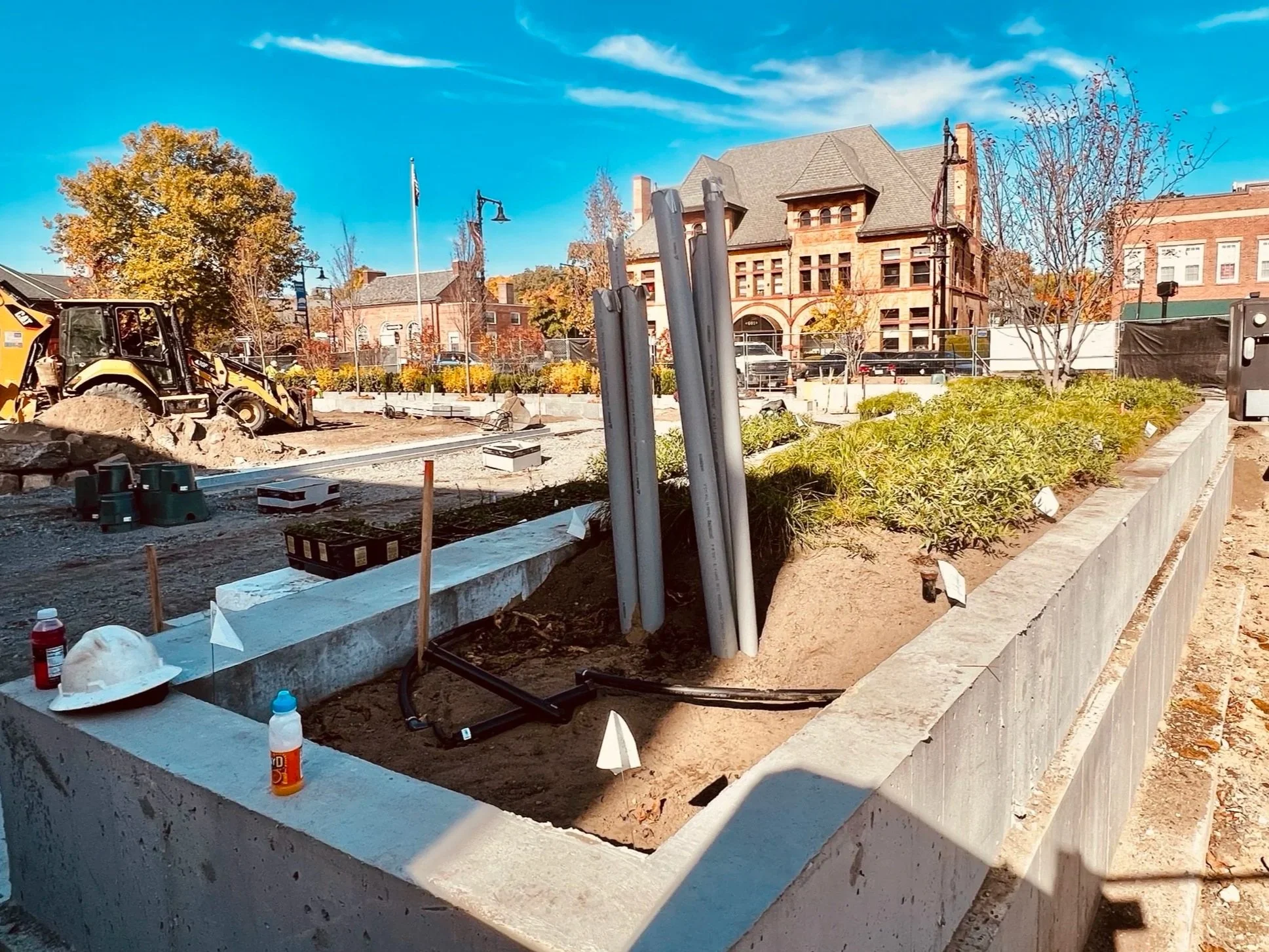 Construction site with trenches, dirt, a backhoe, and pipes, surrounded by a low concrete wall, with trees and historic brick buildings in the background under a bright blue sky.