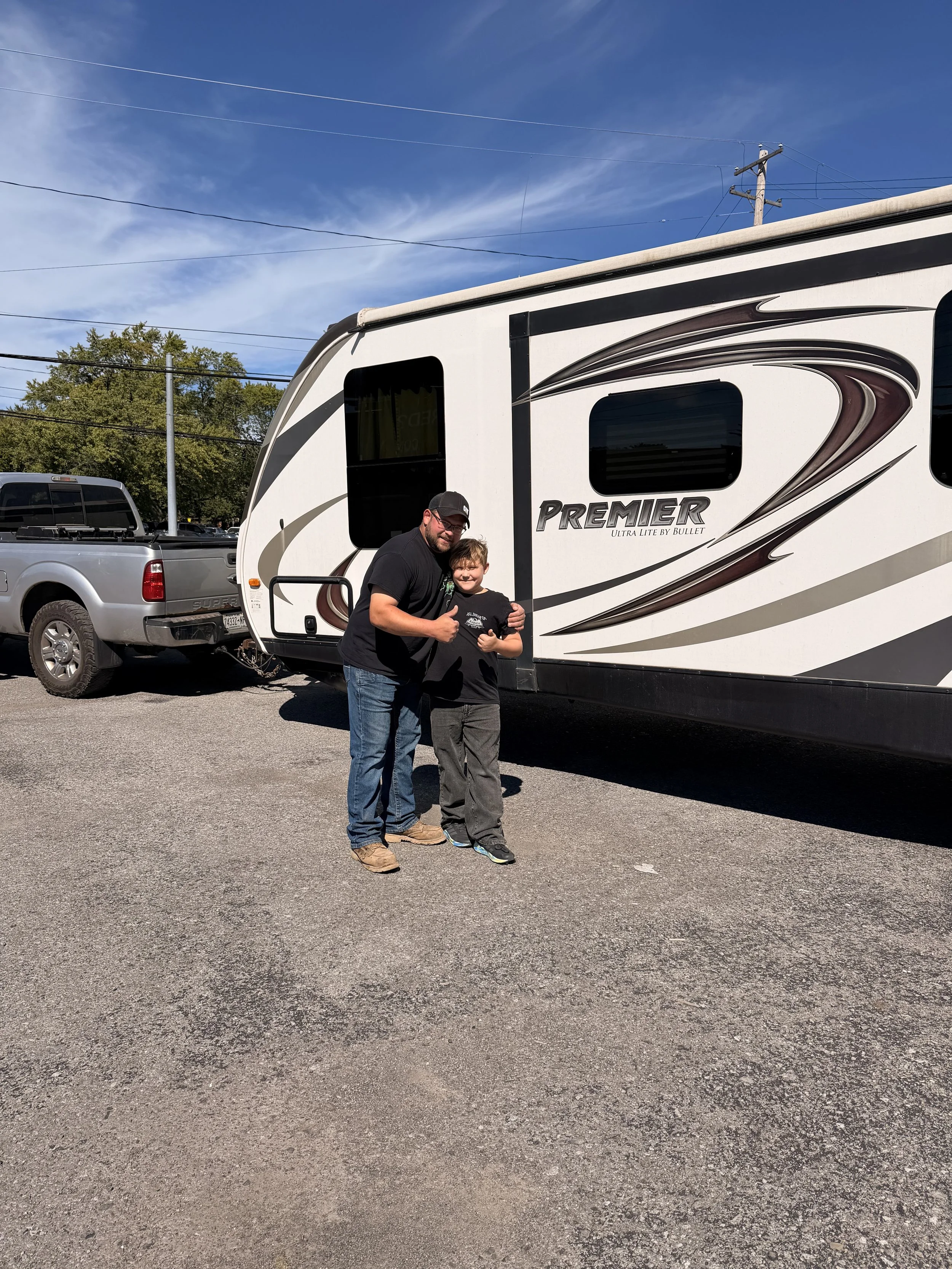 A man and a boy standing next to a white travel trailer, giving a thumbs-up. The trailer is parked in a lot with other vehicles, and there are power lines and trees in the background under a clear blue sky.