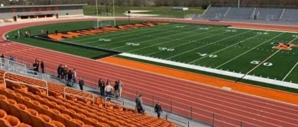 People walking on the track around an empty football field with orange and black colors, in a stadium.