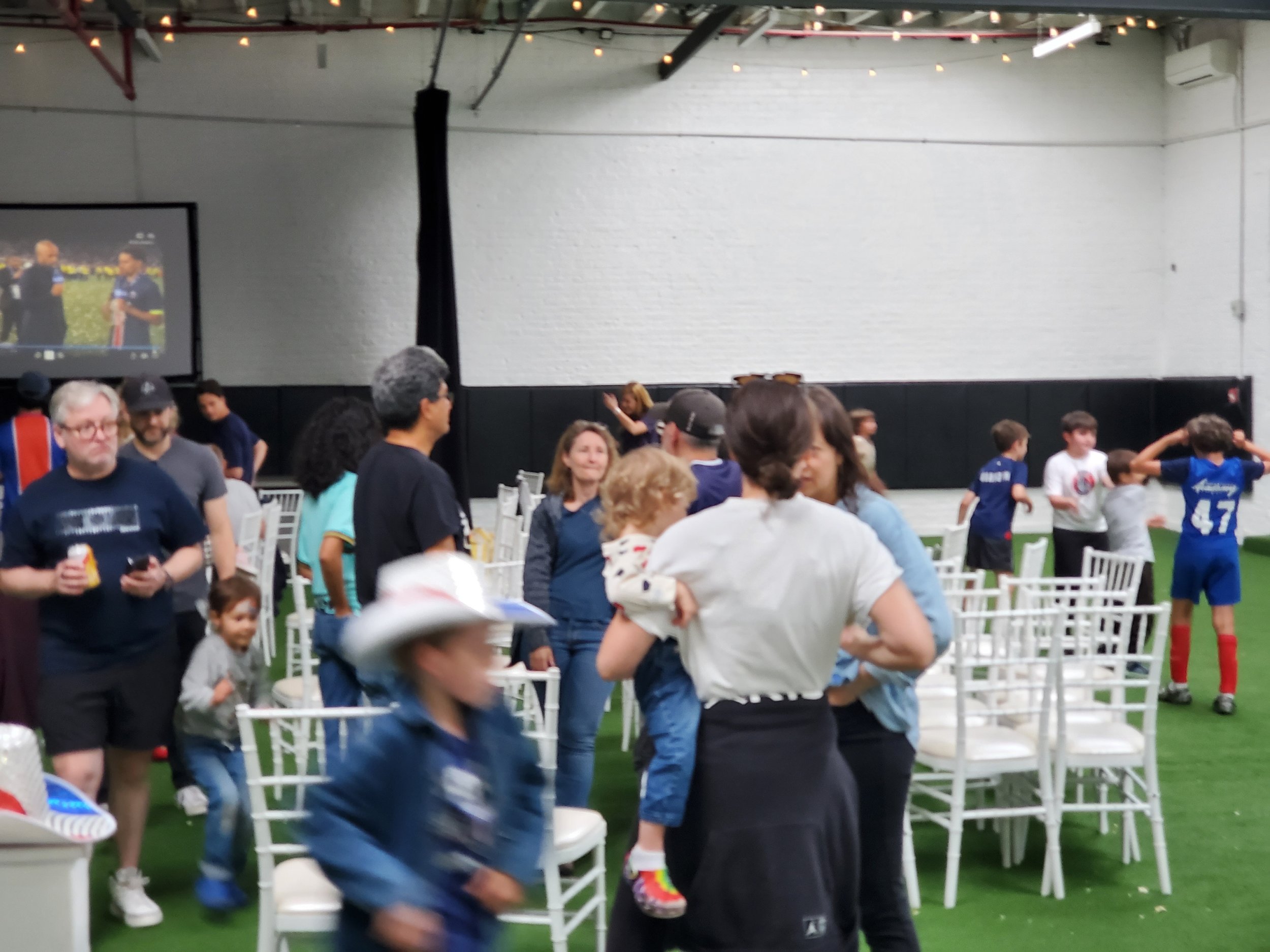 Indoor event with children and adults, some children in sports jerseys, participating in activities and socializing in a facility with white brick walls, string lights, and rows of white chairs.