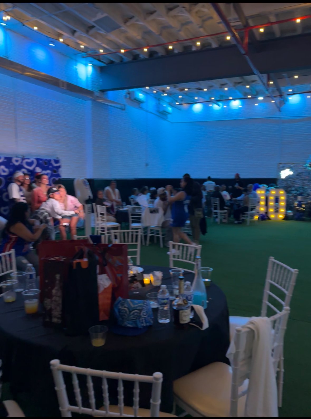 Indoor party setting with round tables, white chairs, and blue lighting. Guests are seated and standing, with a decorated background and large illuminated number 10 in the corner. The tables have drinks and gifts, and the ceiling has string lights.