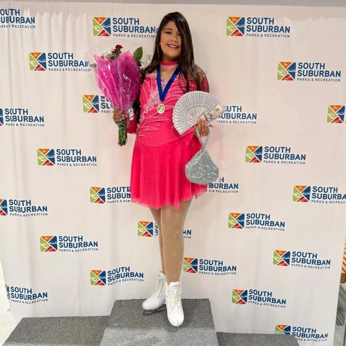 Young girl on ice skates holding a bouquet of pink flowers, a fan, and a sparkly bag, wearing a pink figure skating dress with a medal around her neck, standing on a gray platform in front of a backdrop with the South Suburban Parks & Recreation logo