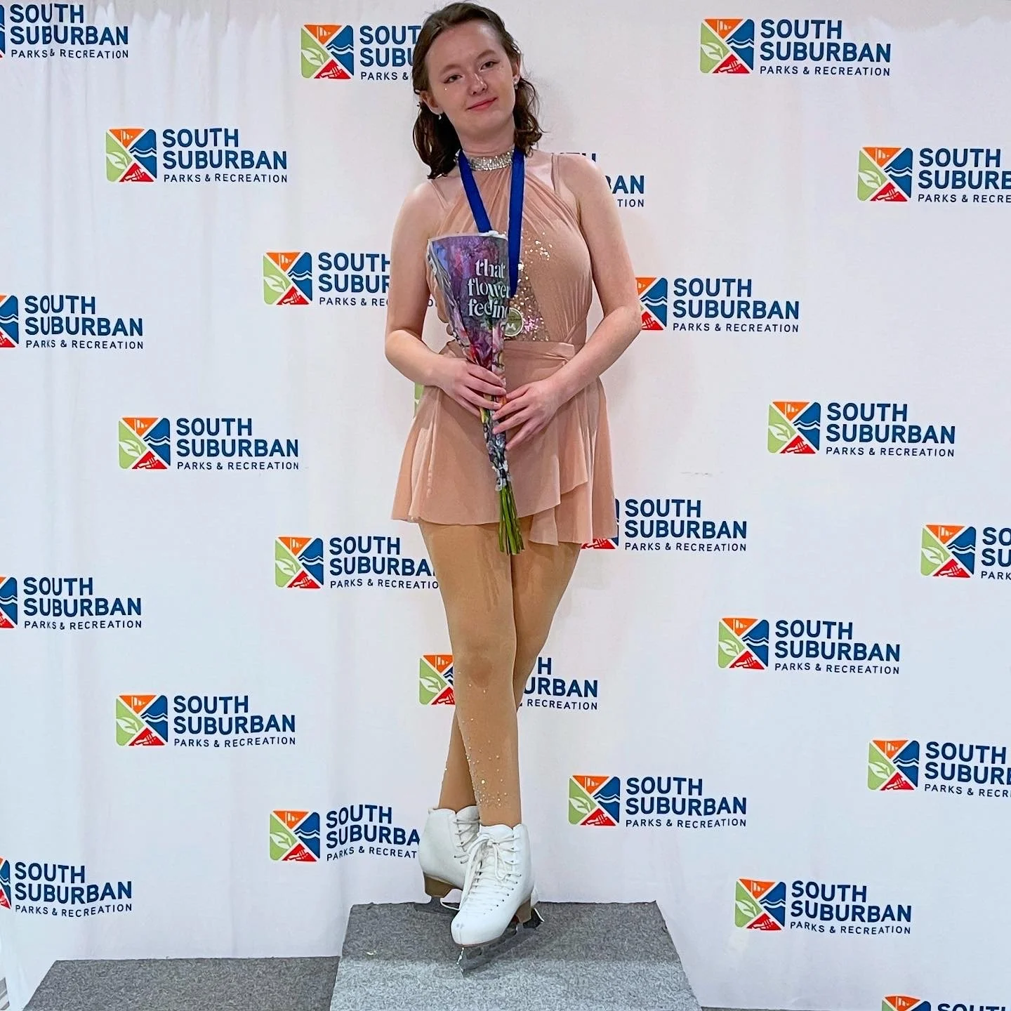A young woman standing on ice skates in front of a backdrop with the logo of South Suburban Parks & Recreation, holding a bouquet of flowers, wearing a peach-colored dress and a silver choker necklace.
