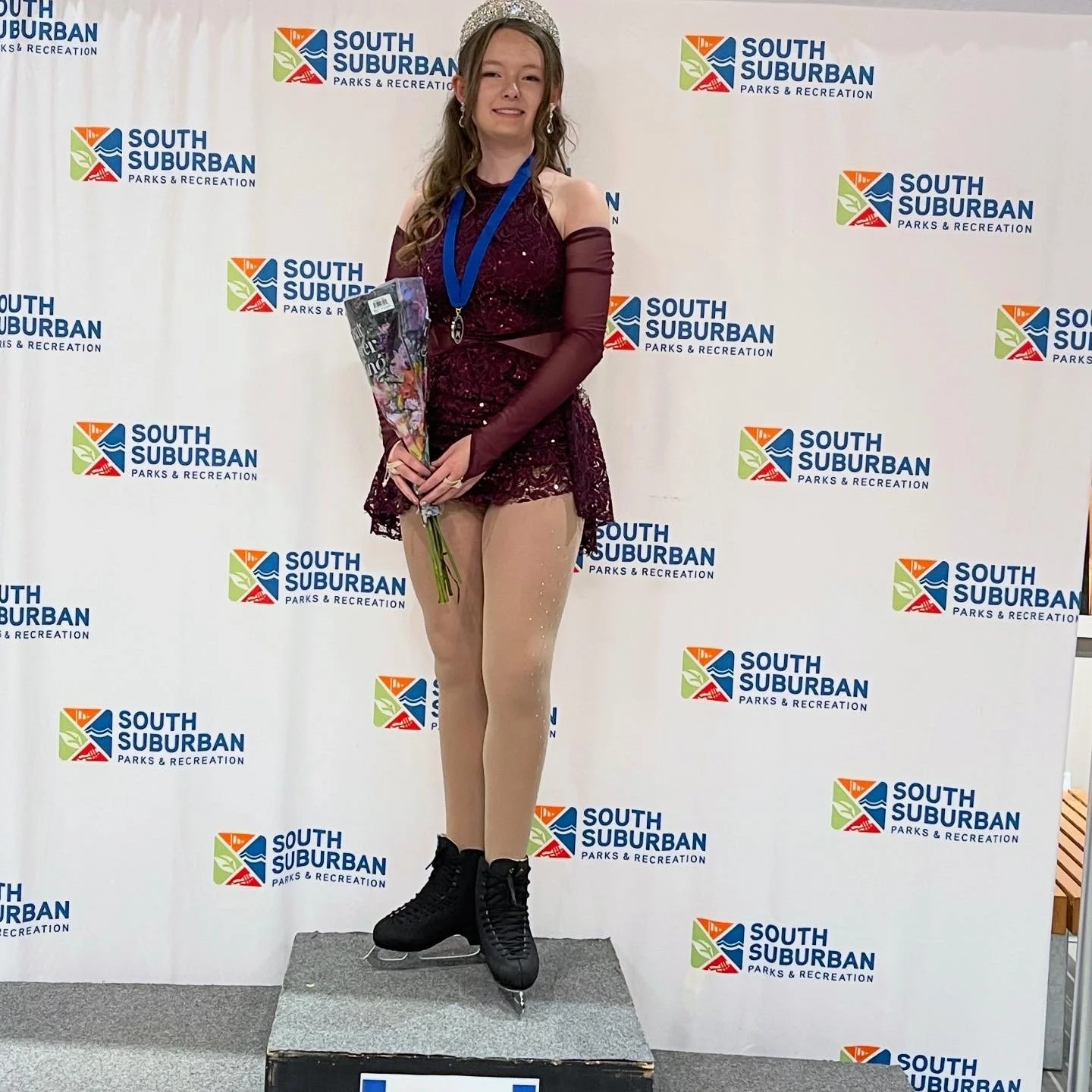 A young girl wearing a tiara and ice skating outfit, holding a bouquet and wearing a medal, standing on a podium with a backdrop featuring the South Suburban Parks & Recreation logo.