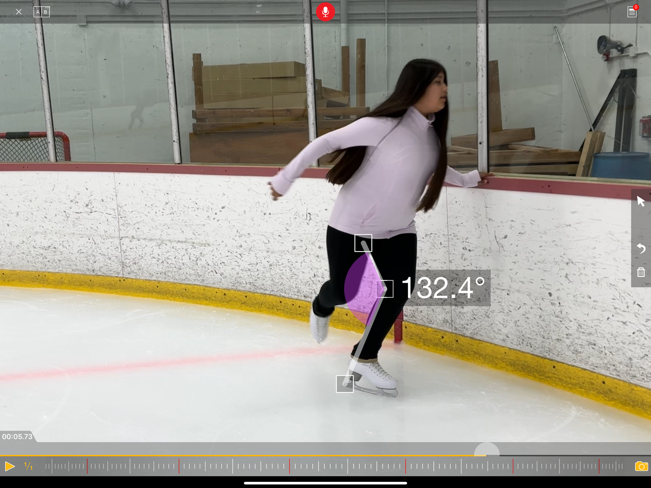 A young woman in a pink hoodie and black pants ice skating inside an ice rink, holding onto the rink's wall for balance, with a pink and purple ice skate blade.