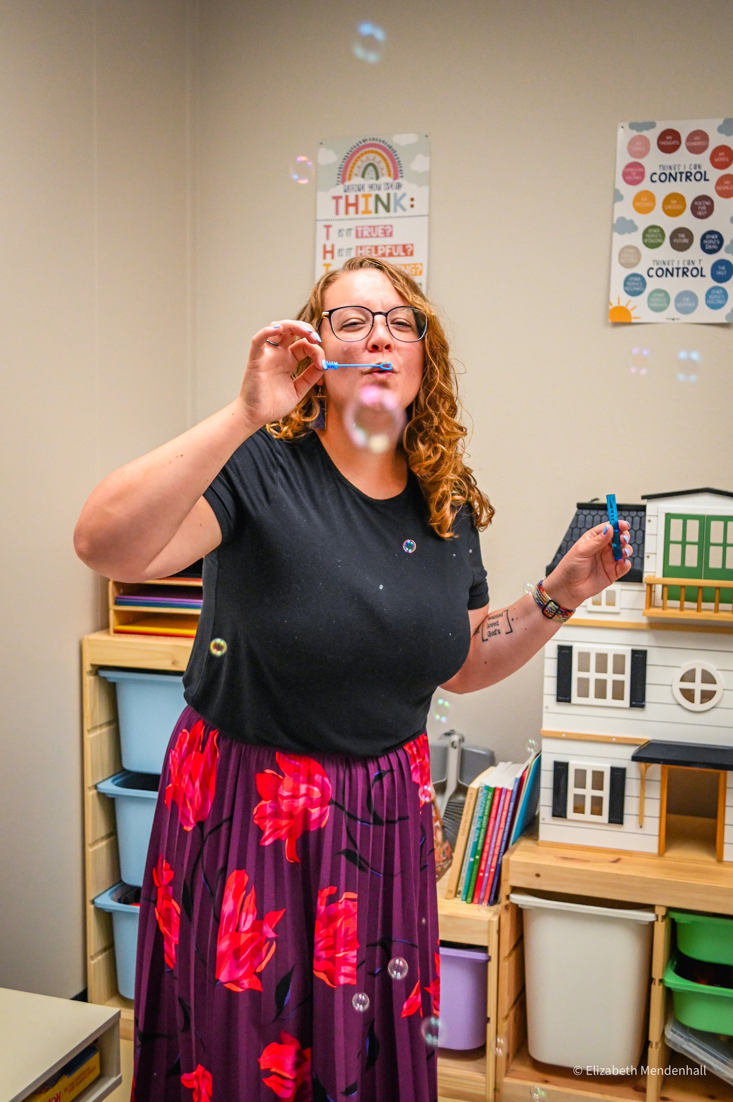 A woman with curly red hair and glasses blowing bubbles in a room with educational posters on the wall and shelves filled with books and storage bins.
